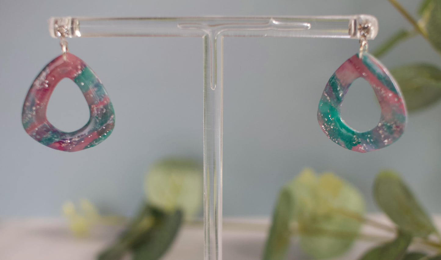 Colorful teardrop-shaped earrings on a clear stand with a blurred green plant background