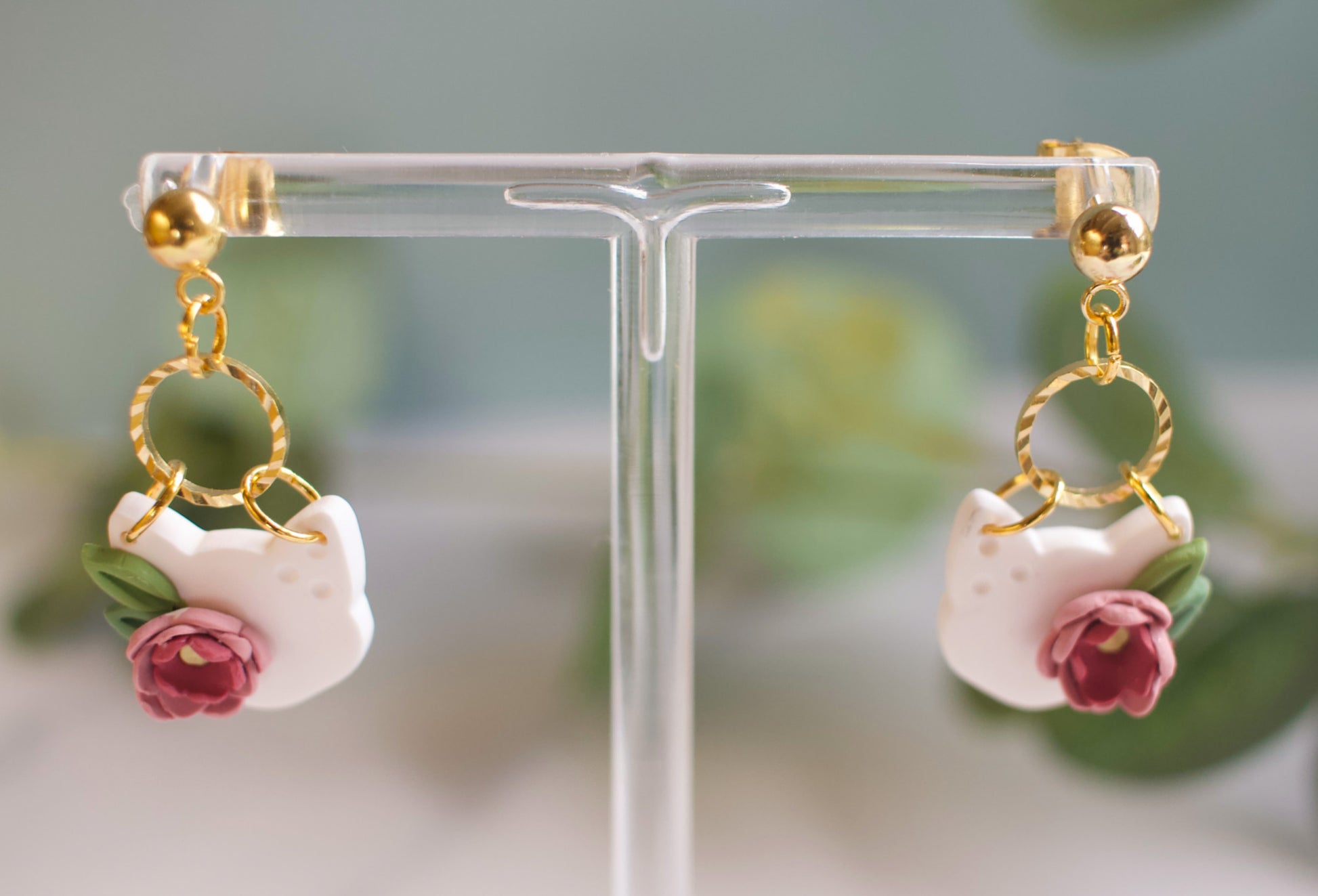 Pair of cat head shaped earrings with floral details on a clear stand.