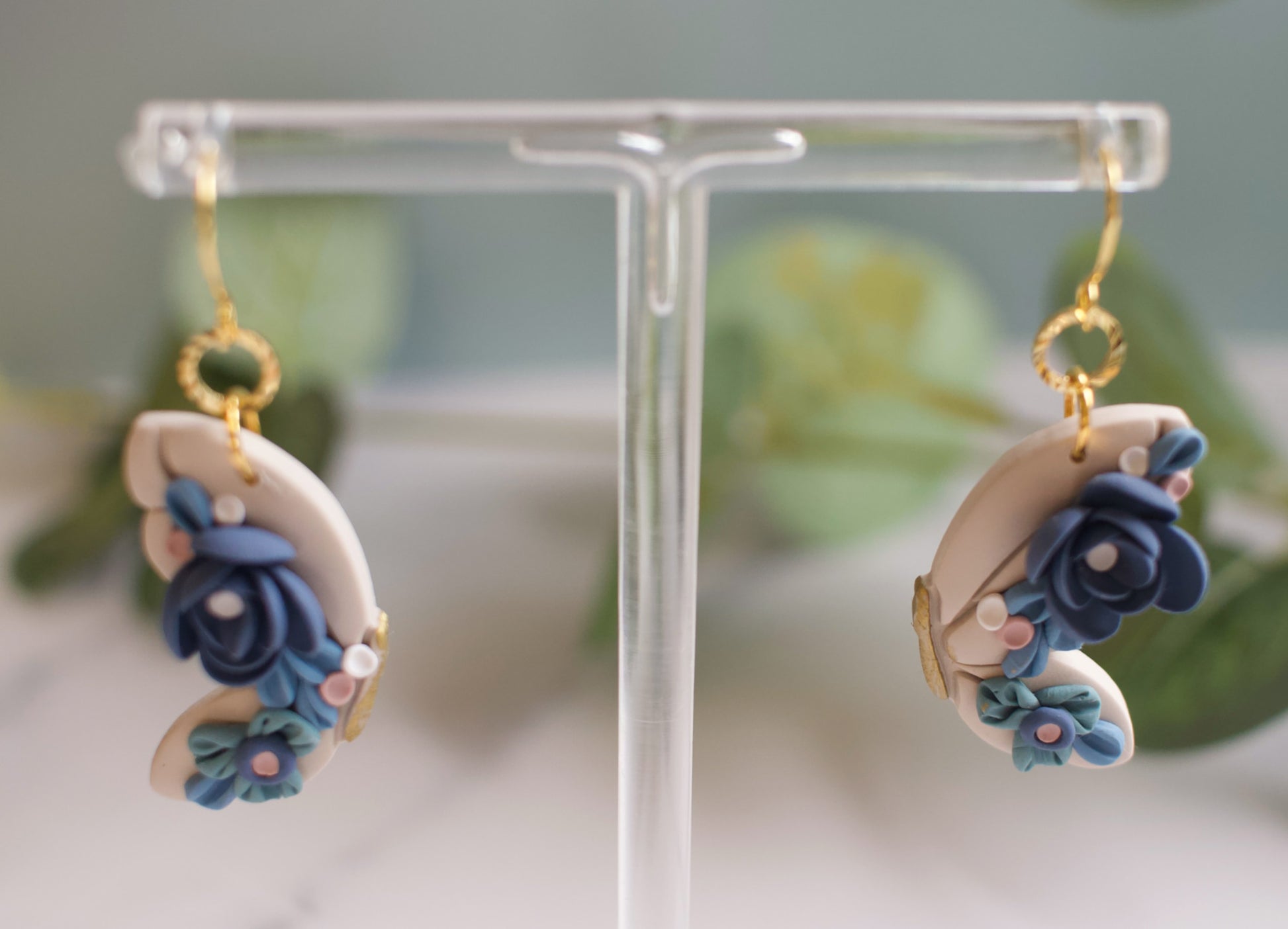 Pair of butterfly wing earrings with blue floral designs on a clear stand against a blurred green background