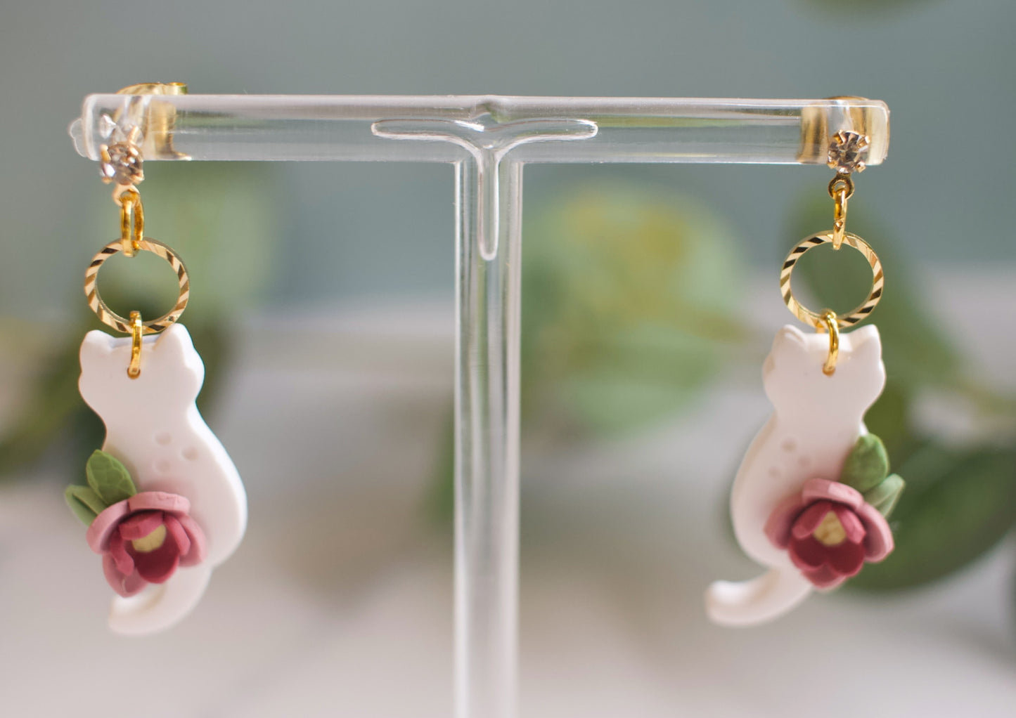 Cat-shaped earrings with floral details on a clear stand against a blurred green background
