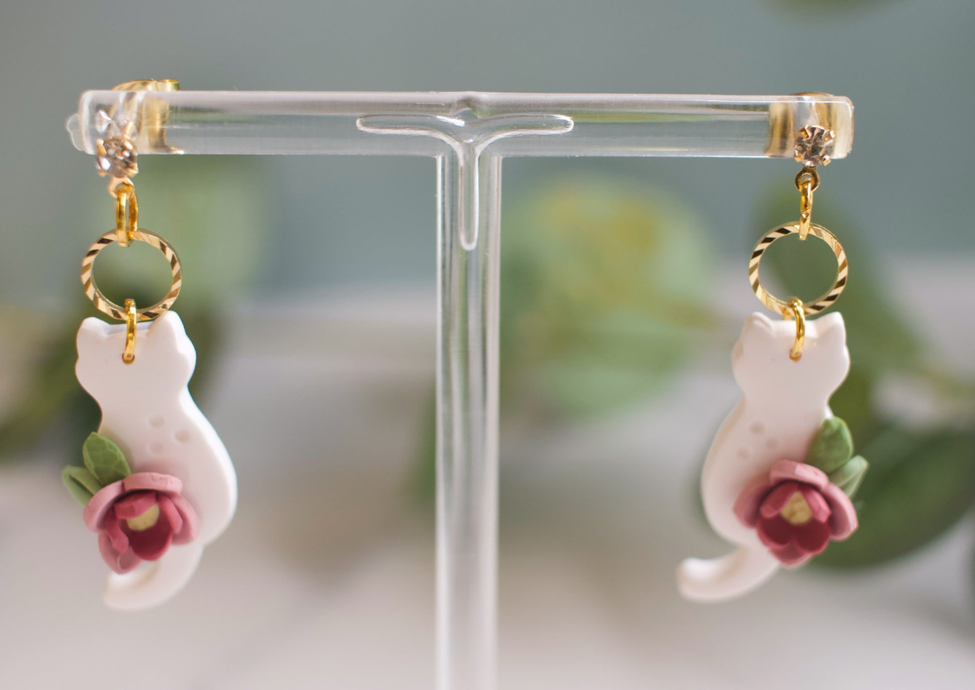 Cat-shaped earrings with floral details on a clear stand against a blurred green background