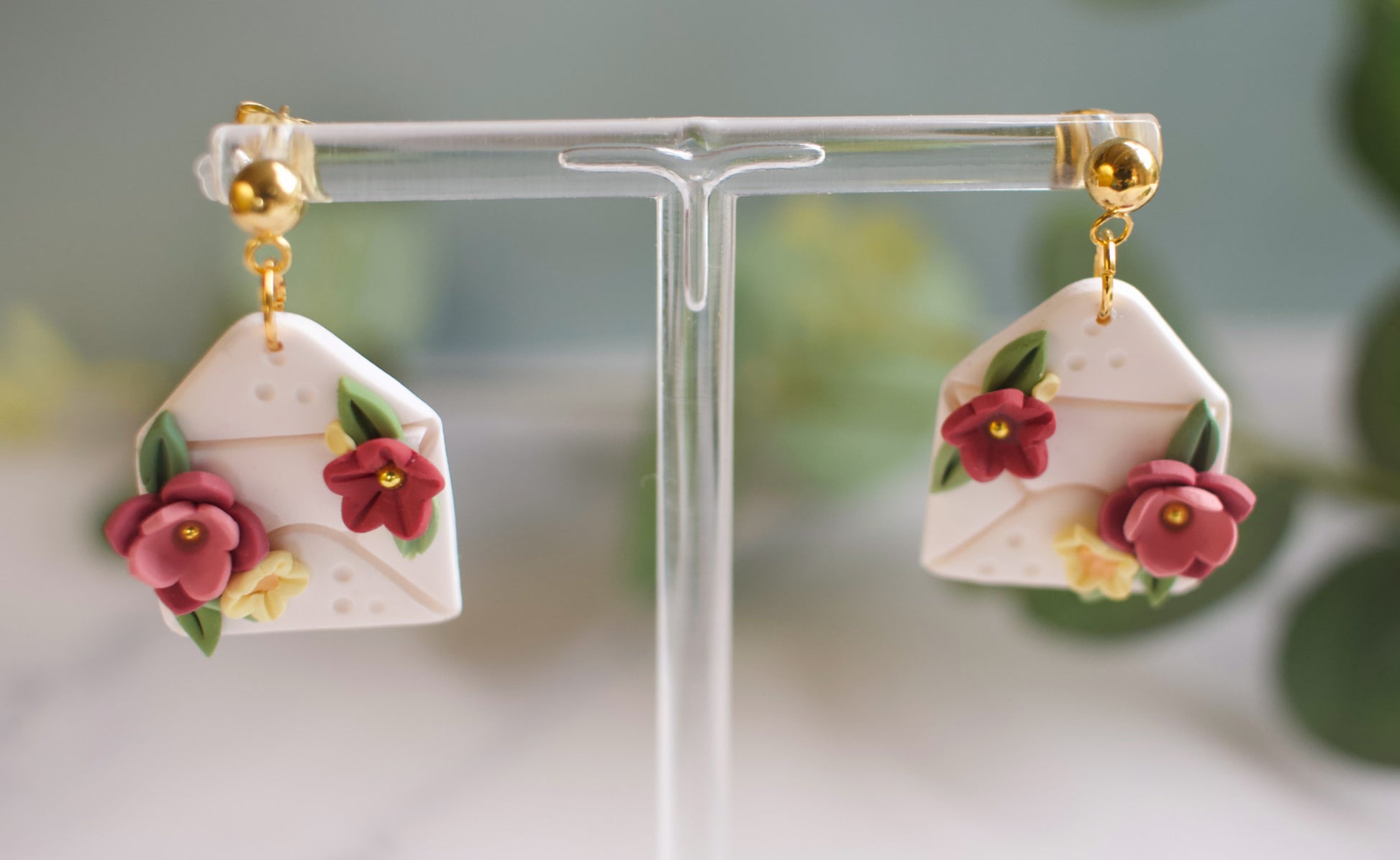 Floral-themed envelope shaped earrings on a clear stand with a blurred green background