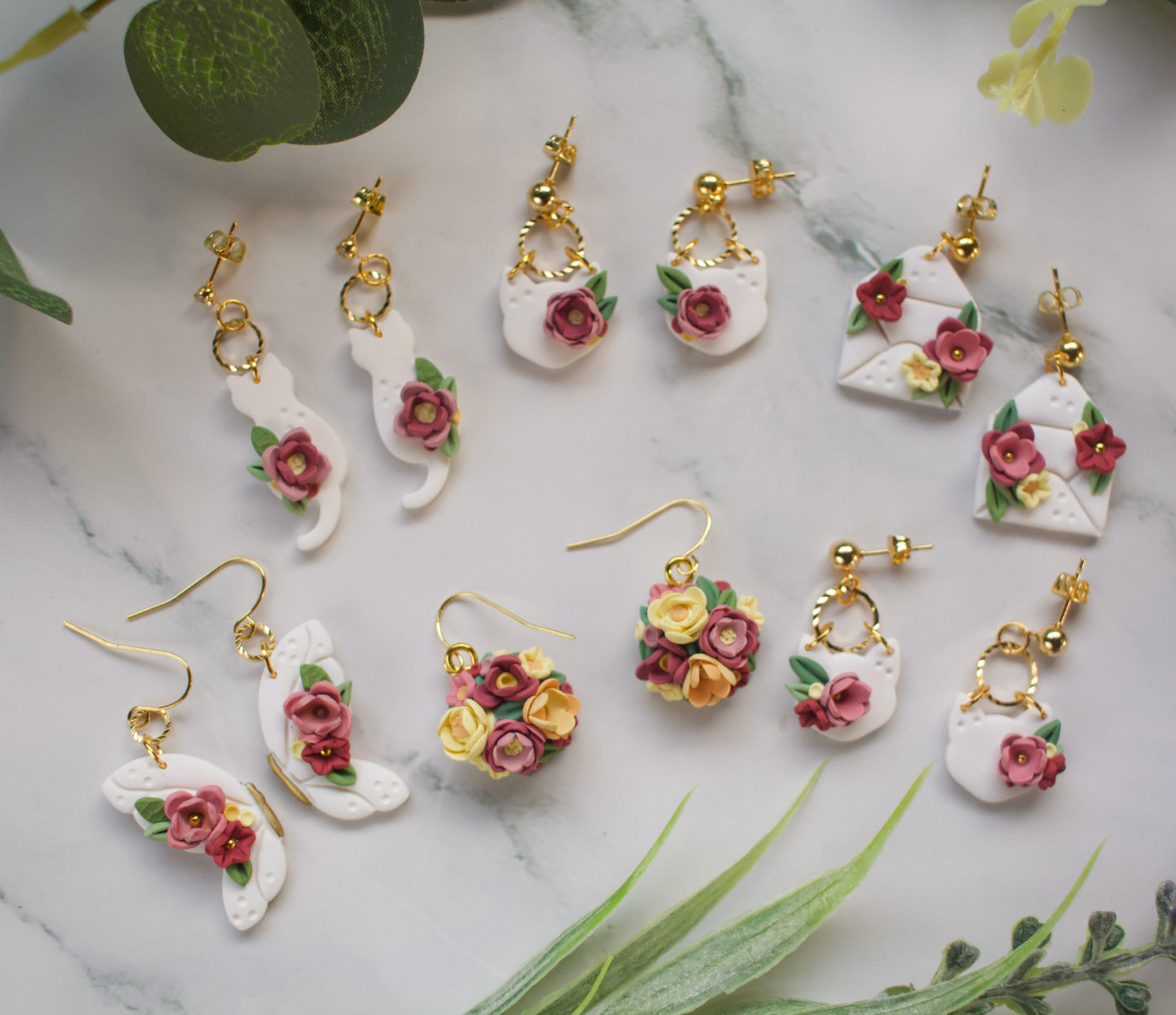 Collection of floral-themed earrings on a marble surface with green leaves.