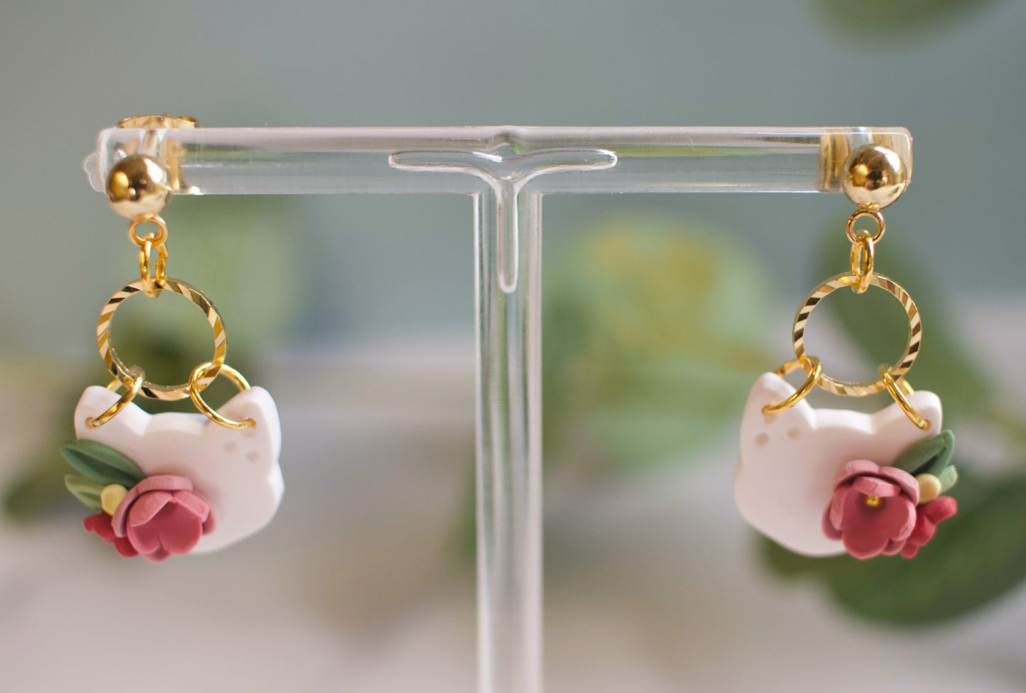 Pair of cat head earrings with gold loops and pink flowers on a clear stand.