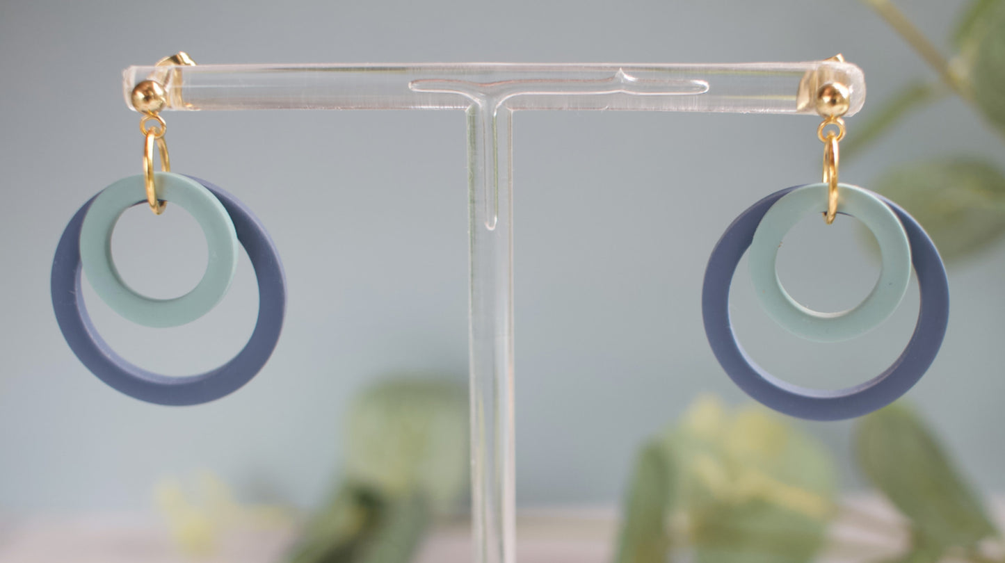 Earrings with blue ad turquoise circular loop design on a clear stand against a blurred natural background