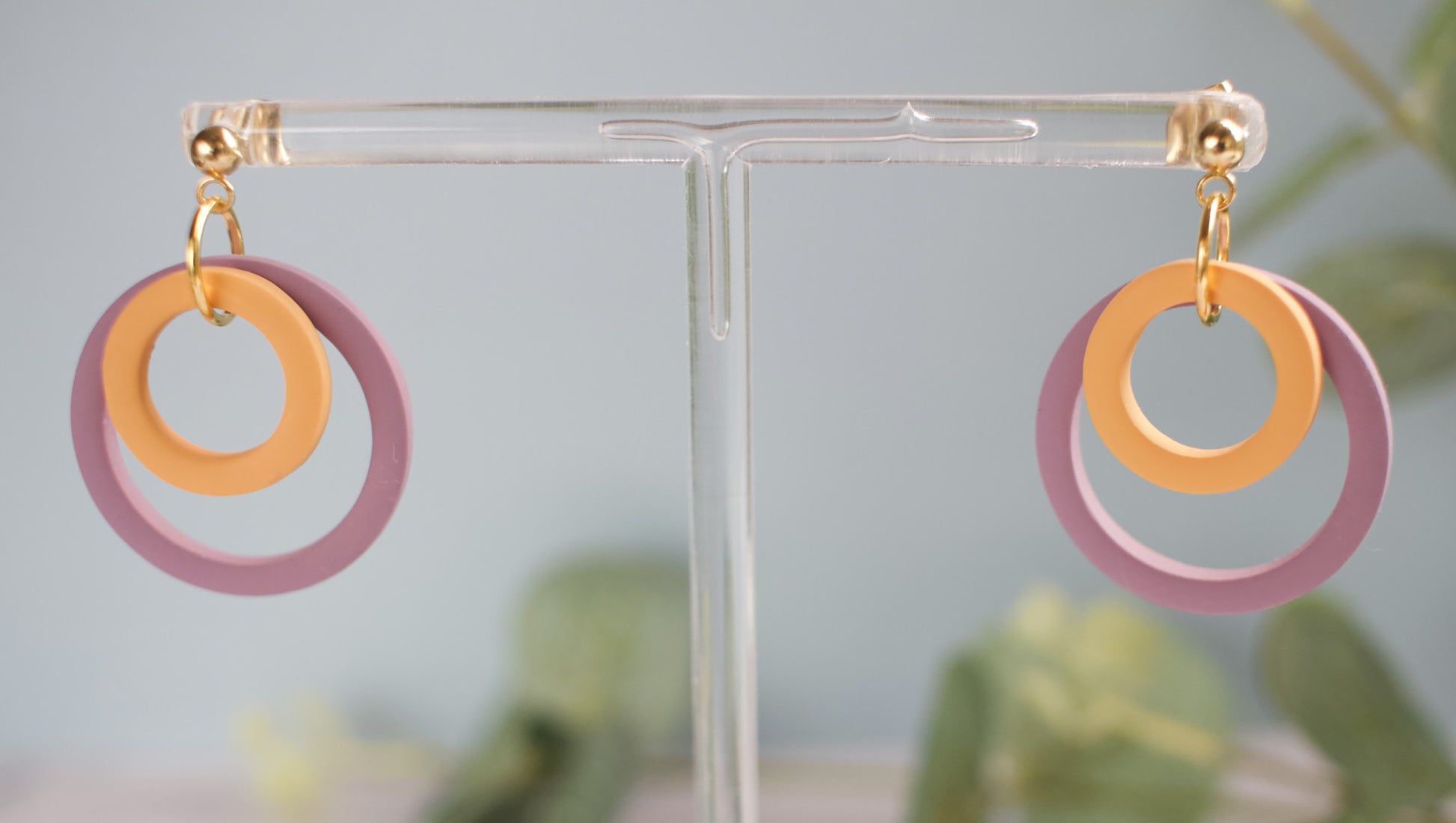 Purple and yellow circular earrings on a clear stand with a blurred green plant background