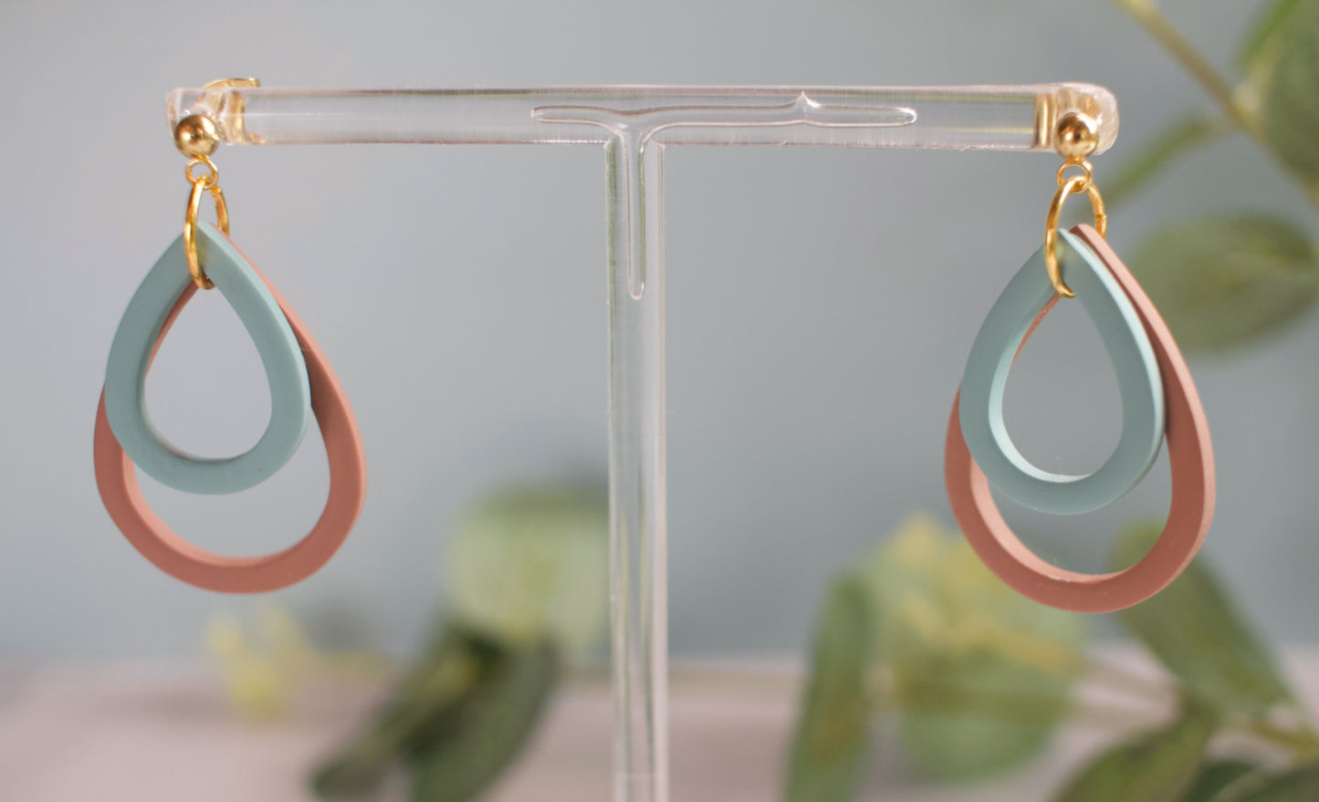 Pair of teal and brown tear drop shaped earrings on a clear stand with a blurred green plant background