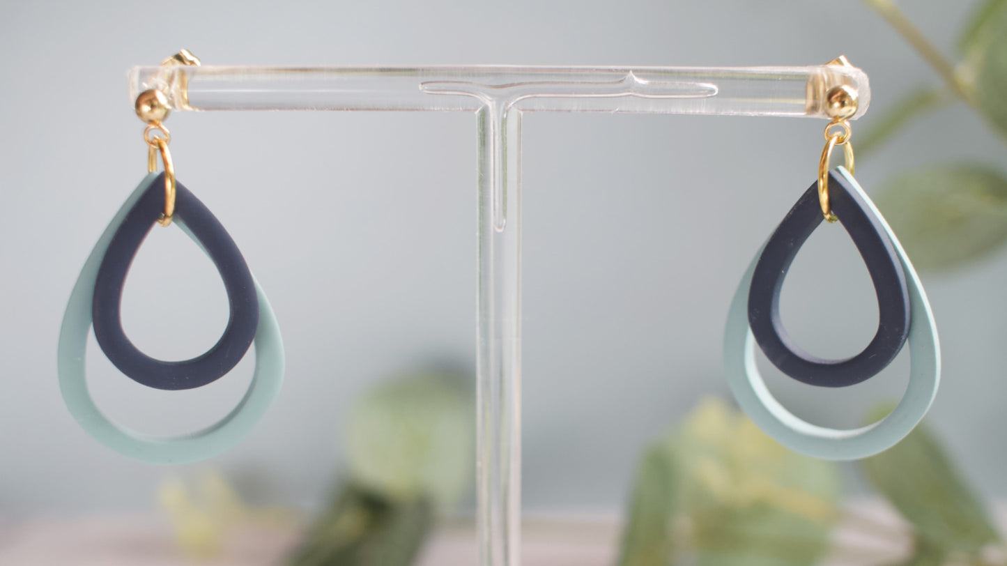 Navy and teal teardrop-shaped earrings on a clear stand with a blurred natural background
