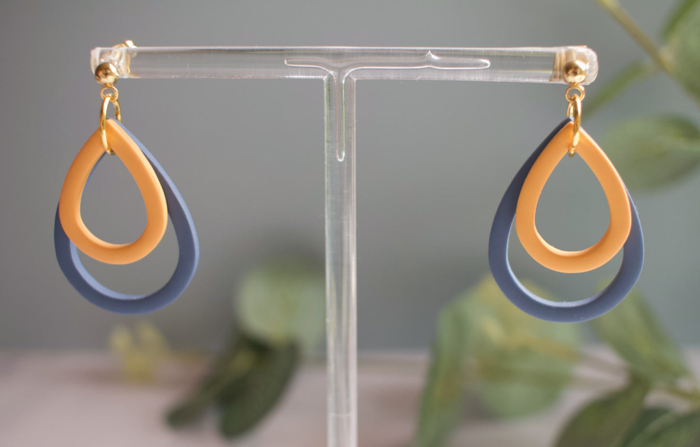 Pair of yellow and blue earrings on a clear stand with a blurred green plant background