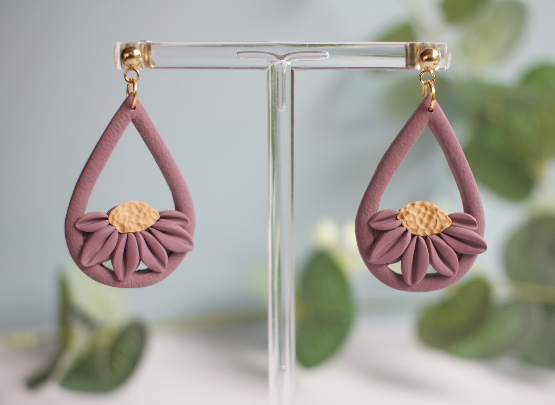 Pair of pink teardrop earrings with gold centers on a clear stand against a blurred green leafy background.