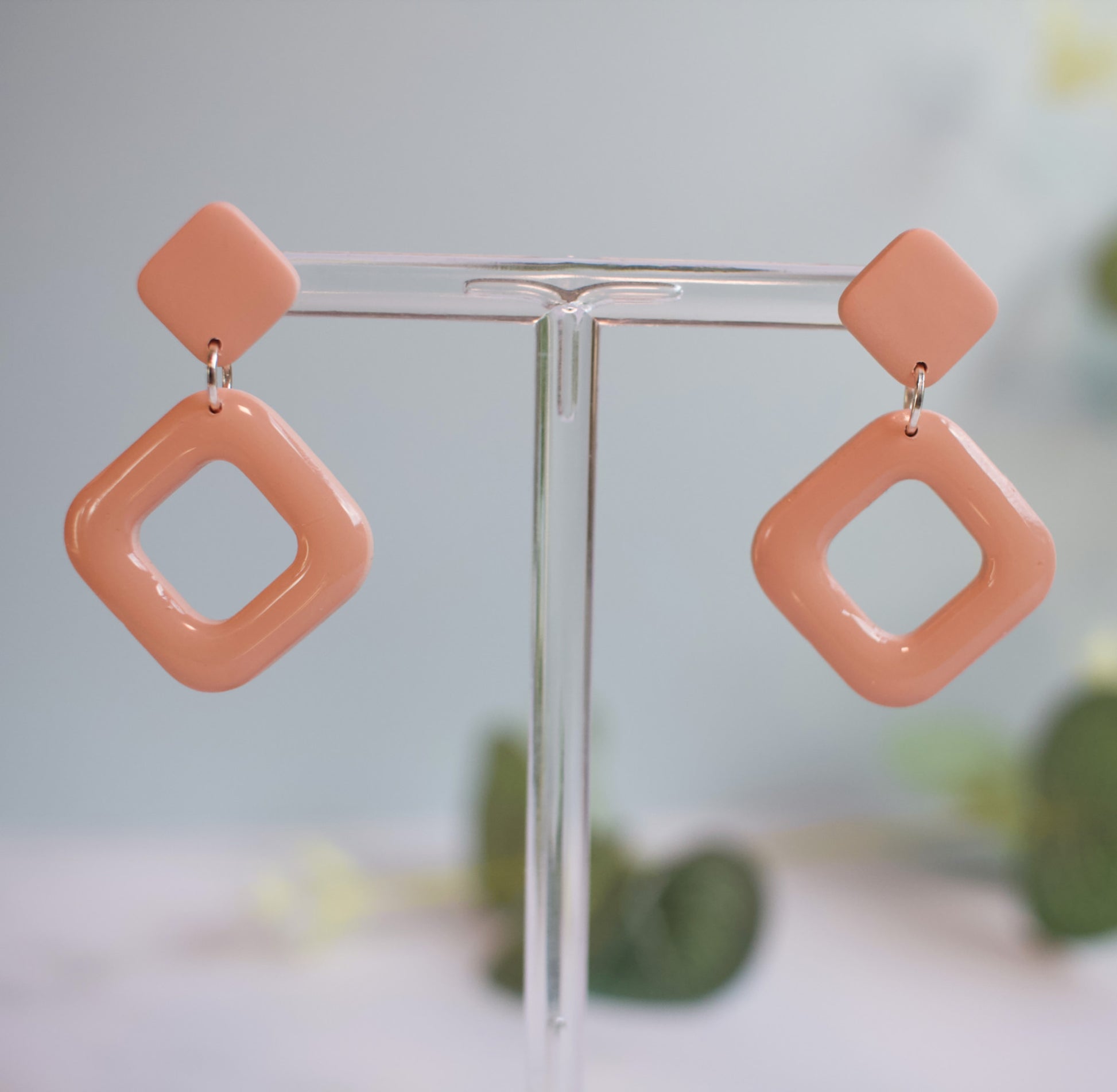 Pair of peach-colored square-shaped earrings on a clear stand with a blurred green background