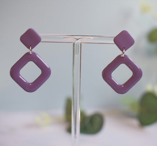 Purple square-shaped earrings on a clear stand with a blurred greenery background