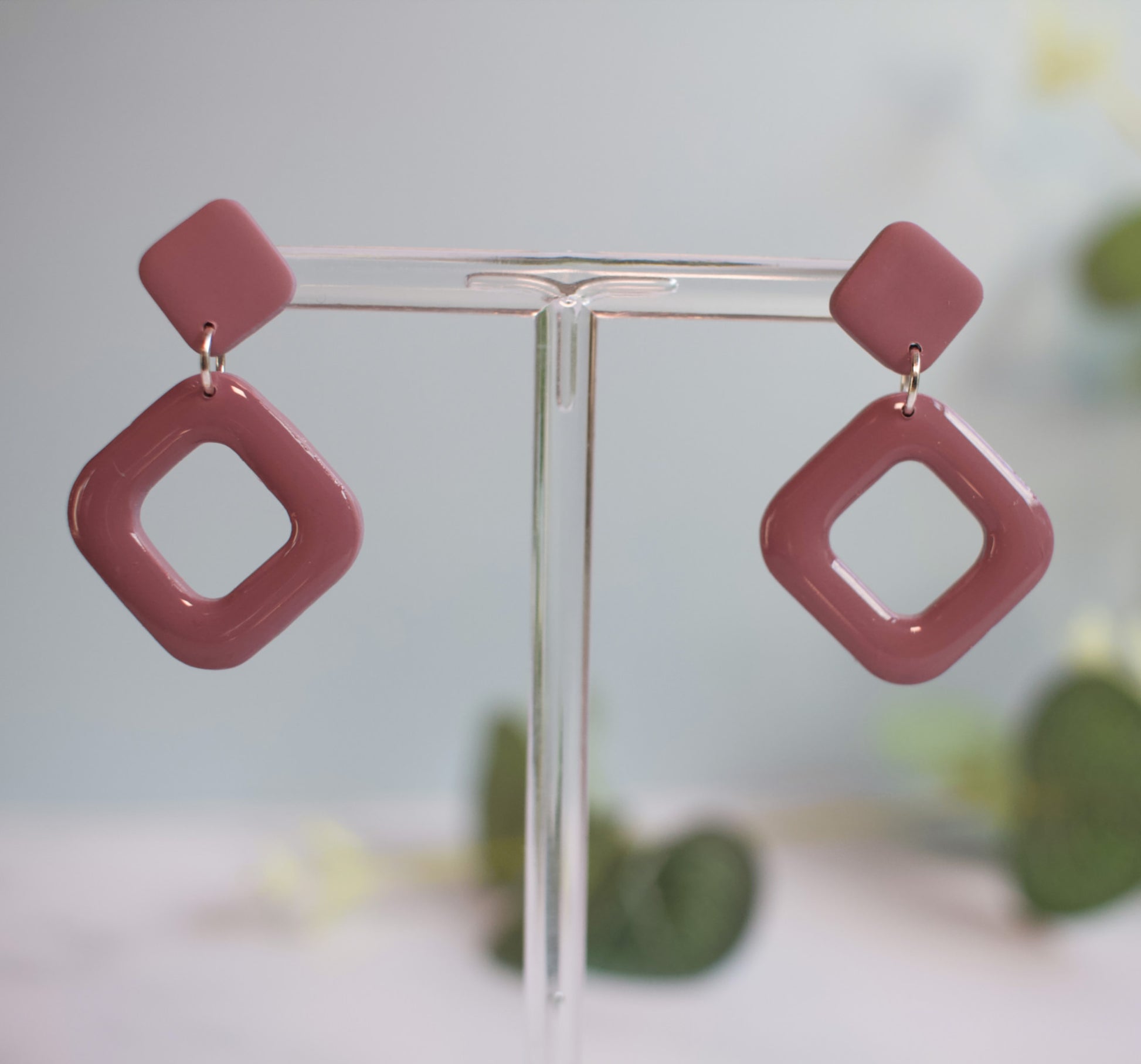 Mulberry coloured square-shaped earrings on a clear stand with a blurred green and yellow background