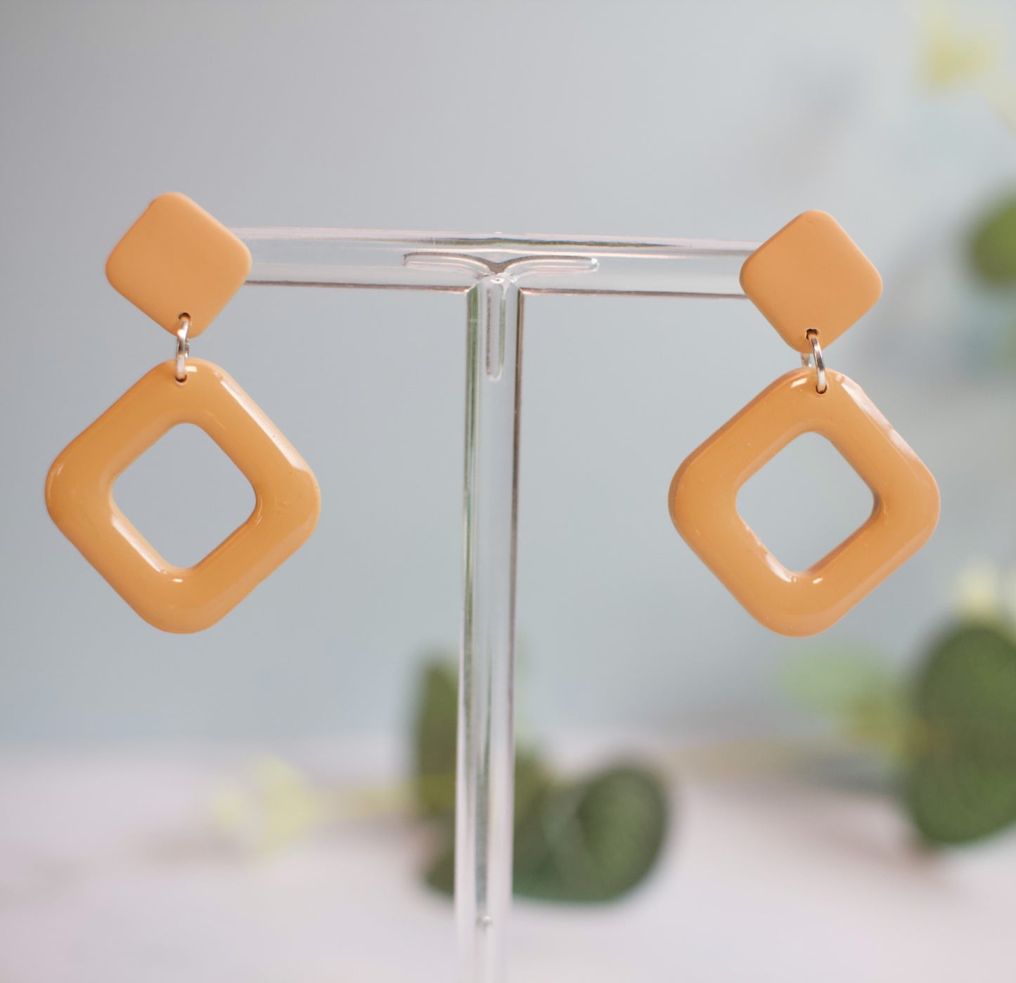 Pair of yellow square-shaped earrings on a clear stand with a blurred green plant background