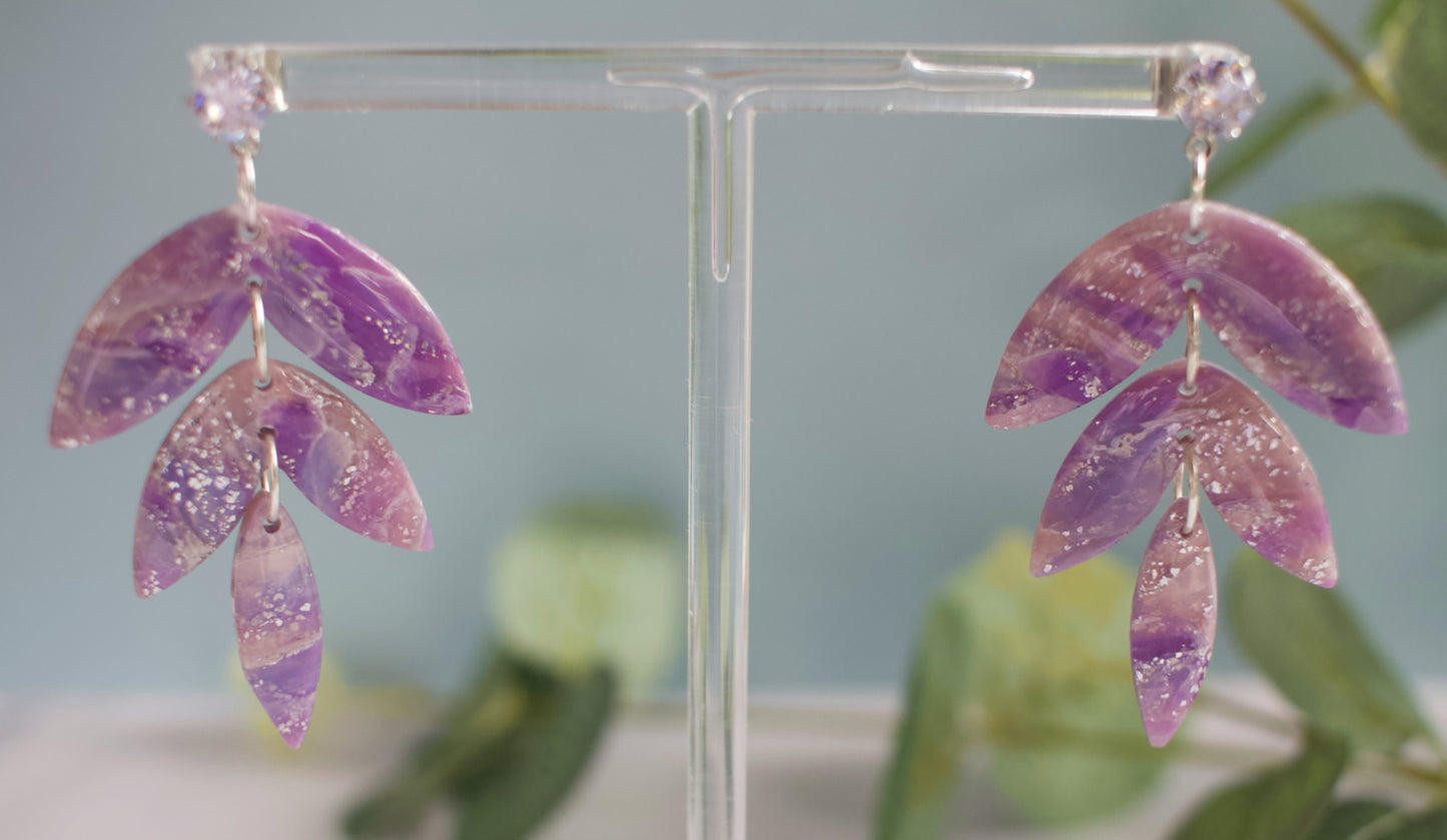 Purple leaf-shaped dangle earrings on a clear stand with a blurred green background