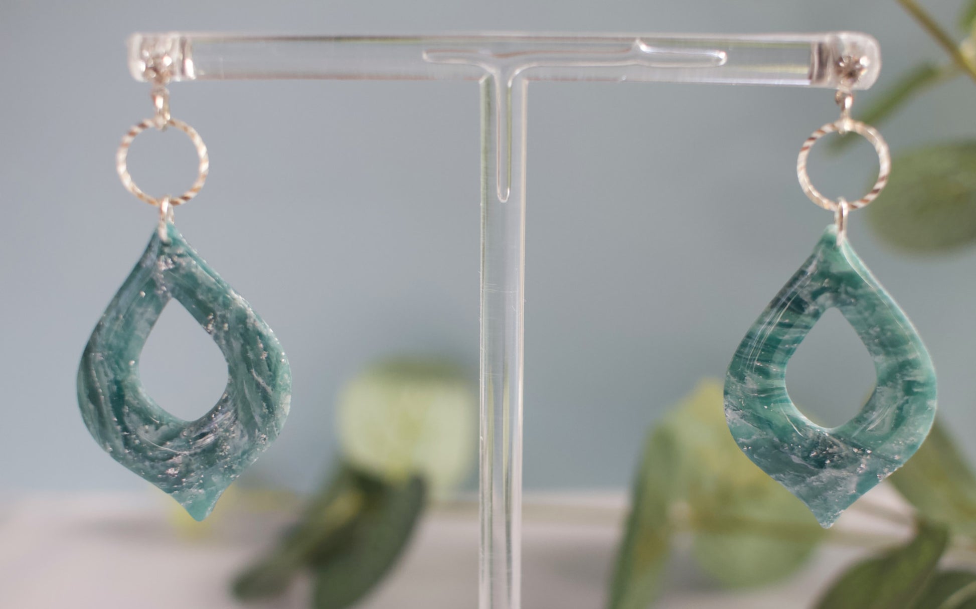 Green teardrop earrings on a clear stand with a blurred green plant background