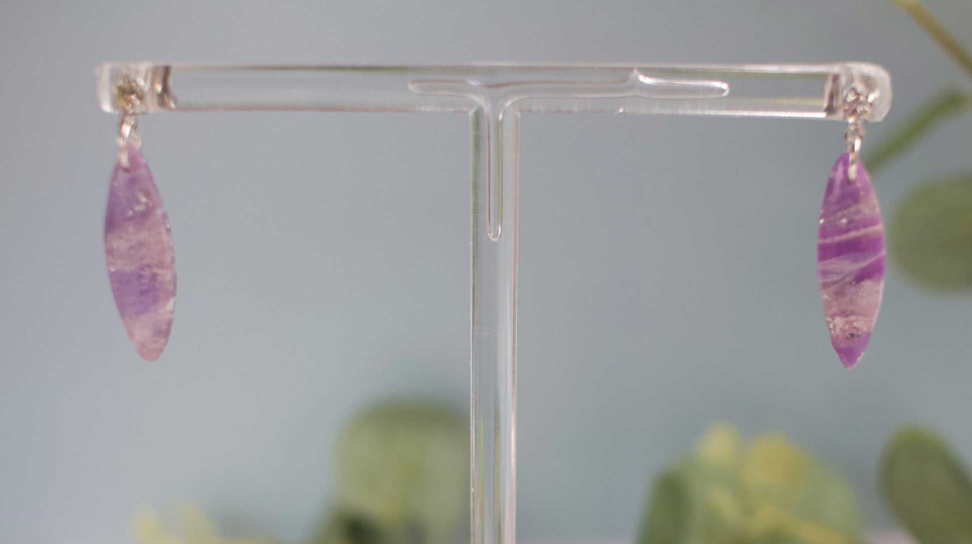 Purple teardrop earrings on a clear stand with a blurred green plant background