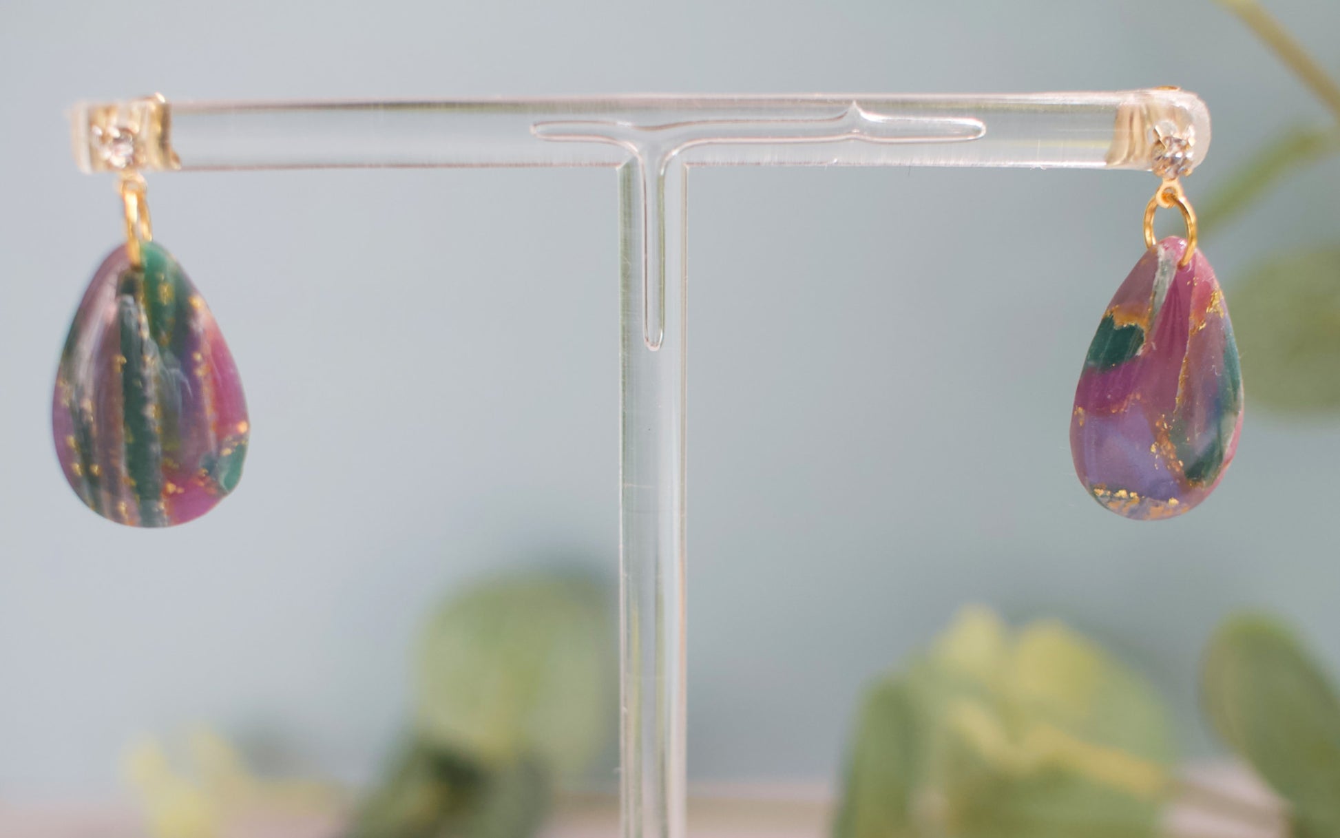 Purple and teal teardrop-shaped earrings on a clear stand with a blurred green background