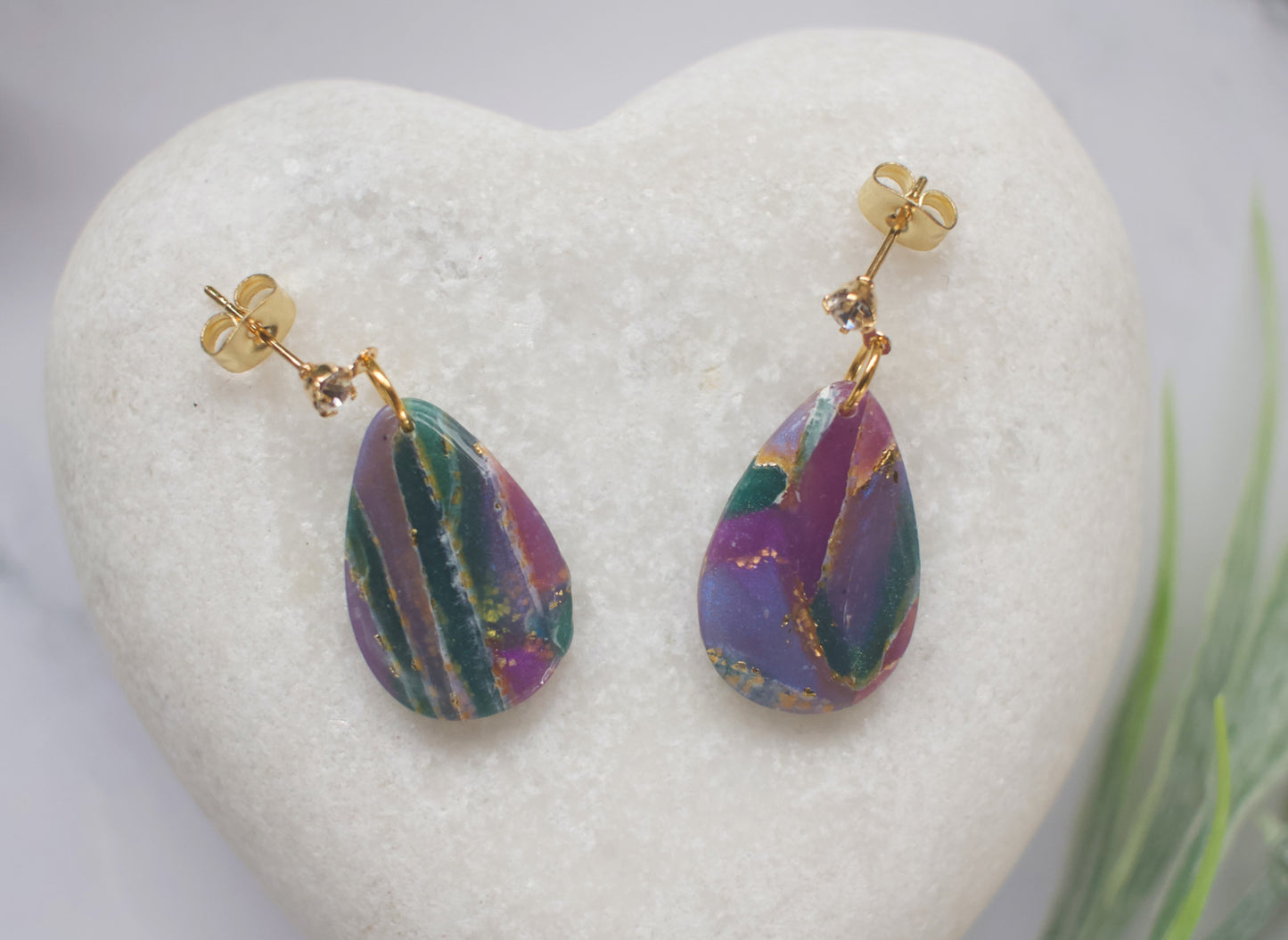 Purple and teal teardrop earrings on a white heart-shaped stand with a blurred green plant in the background.