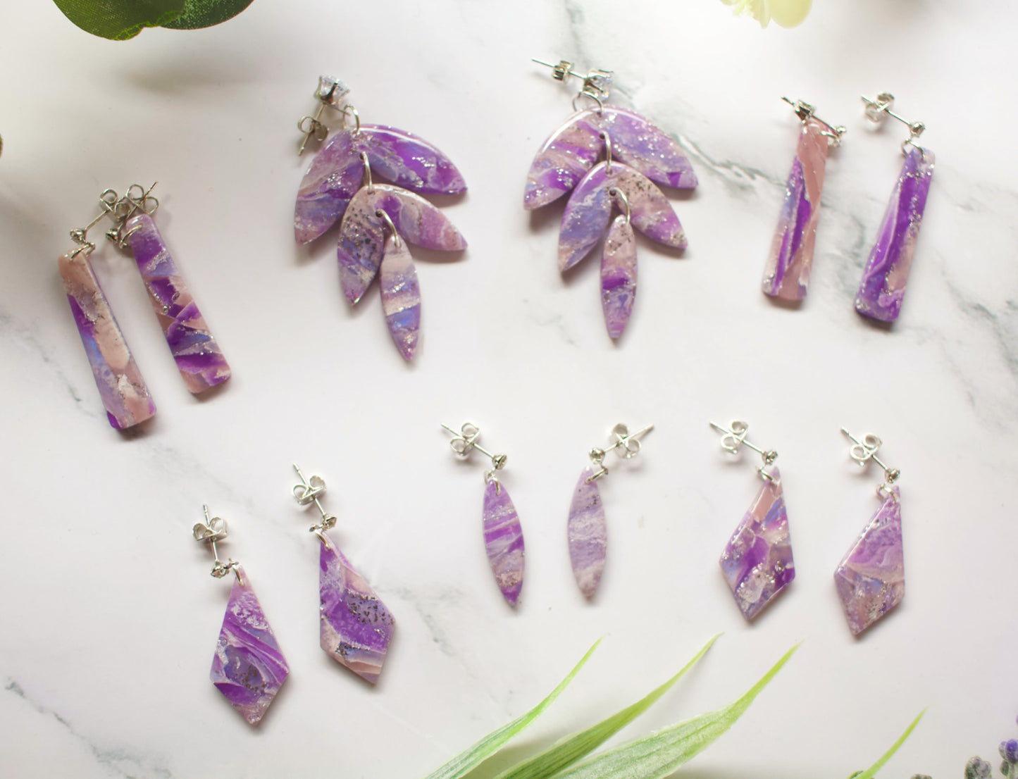 Set of purple and pink marbled earrings on a marble surface with green leaves.