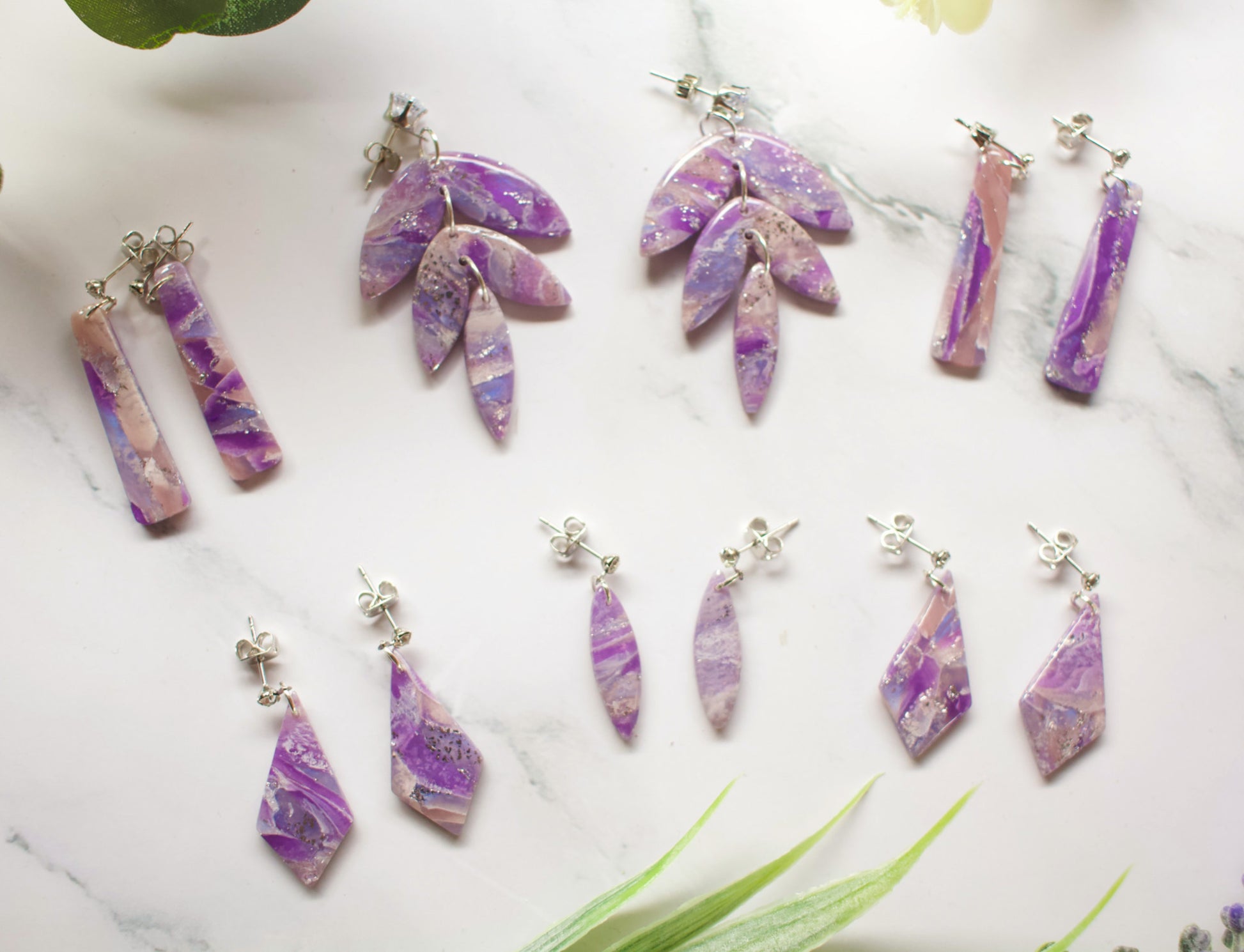 Set of purple and pink marbled earrings on a marble surface with green leaves.