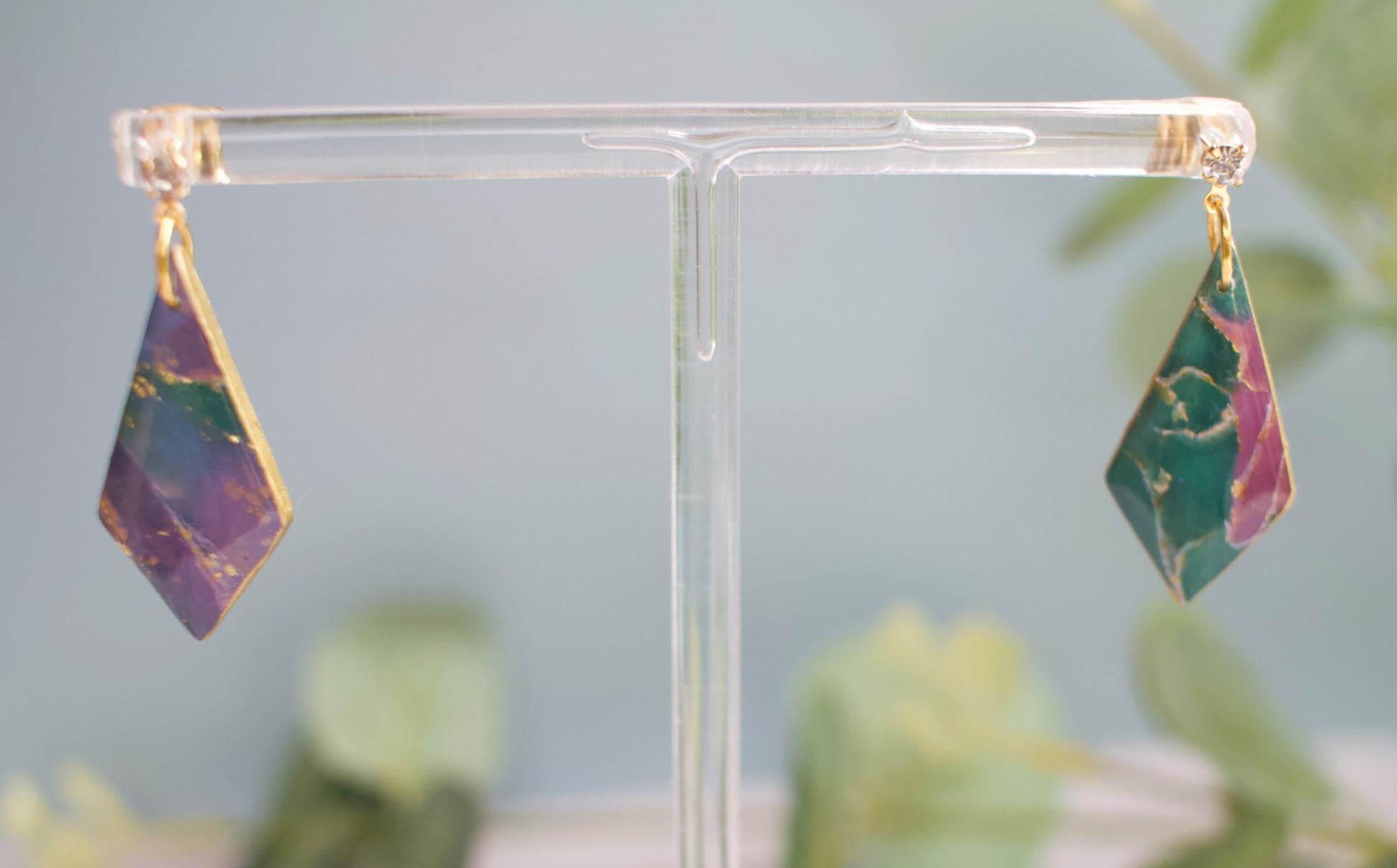 Pair of purple and teal earrings on a clear stand with a blurred green plant background