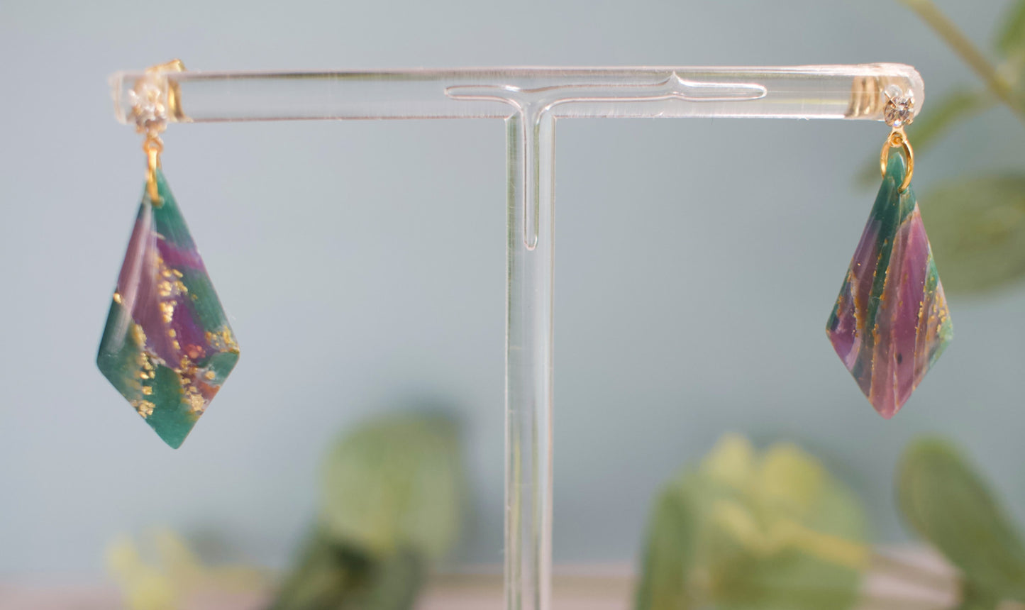 Teal and purple teardrop earrings on a clear stand with a blurred green plant background