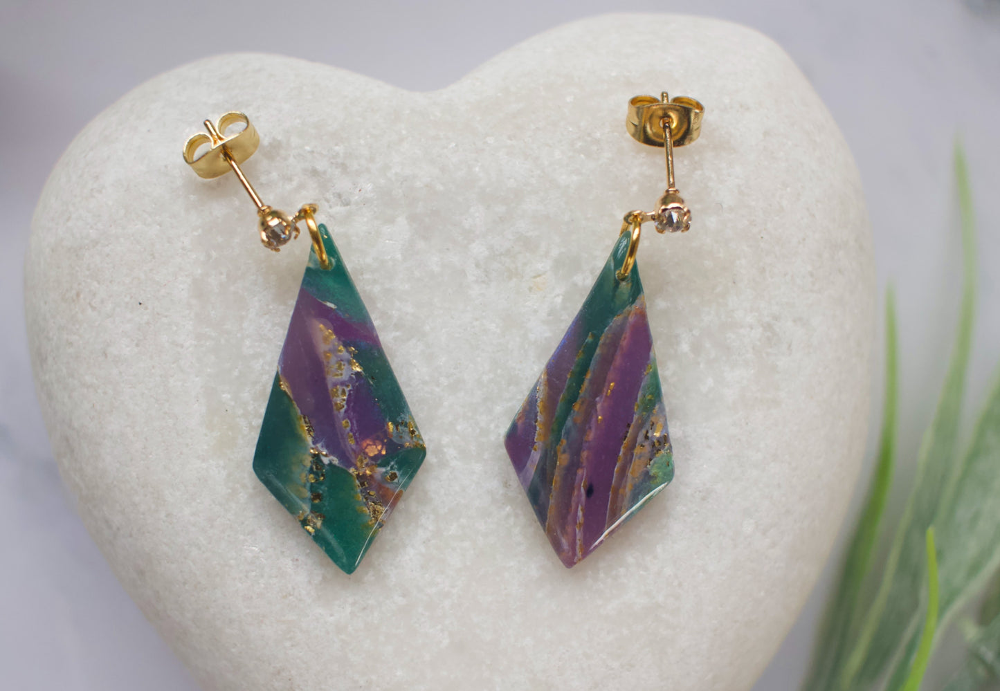Pair of purple and teal marbled diamond-shaped earrings with gold hooks on a heart-shaped stone.