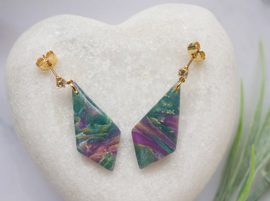 Purple and teal marbled geometric earrings on a heart-shaped stone with a blurred green plant in the background