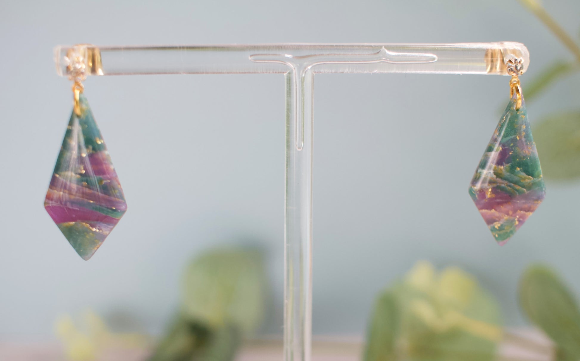 Purple and teal geometric earrings on a clear stand with a blurred green plant background