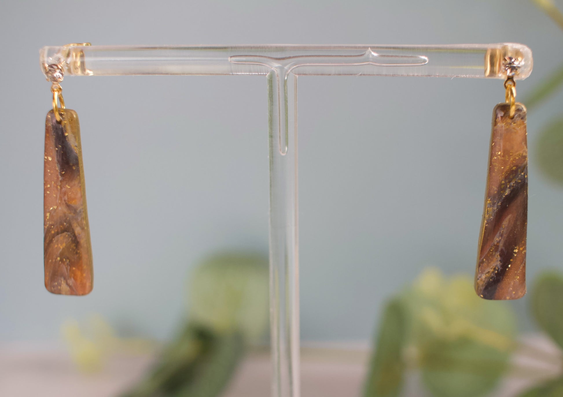 Pair of brown and black marbled long drop earrings on a clear stand with a blurred green background