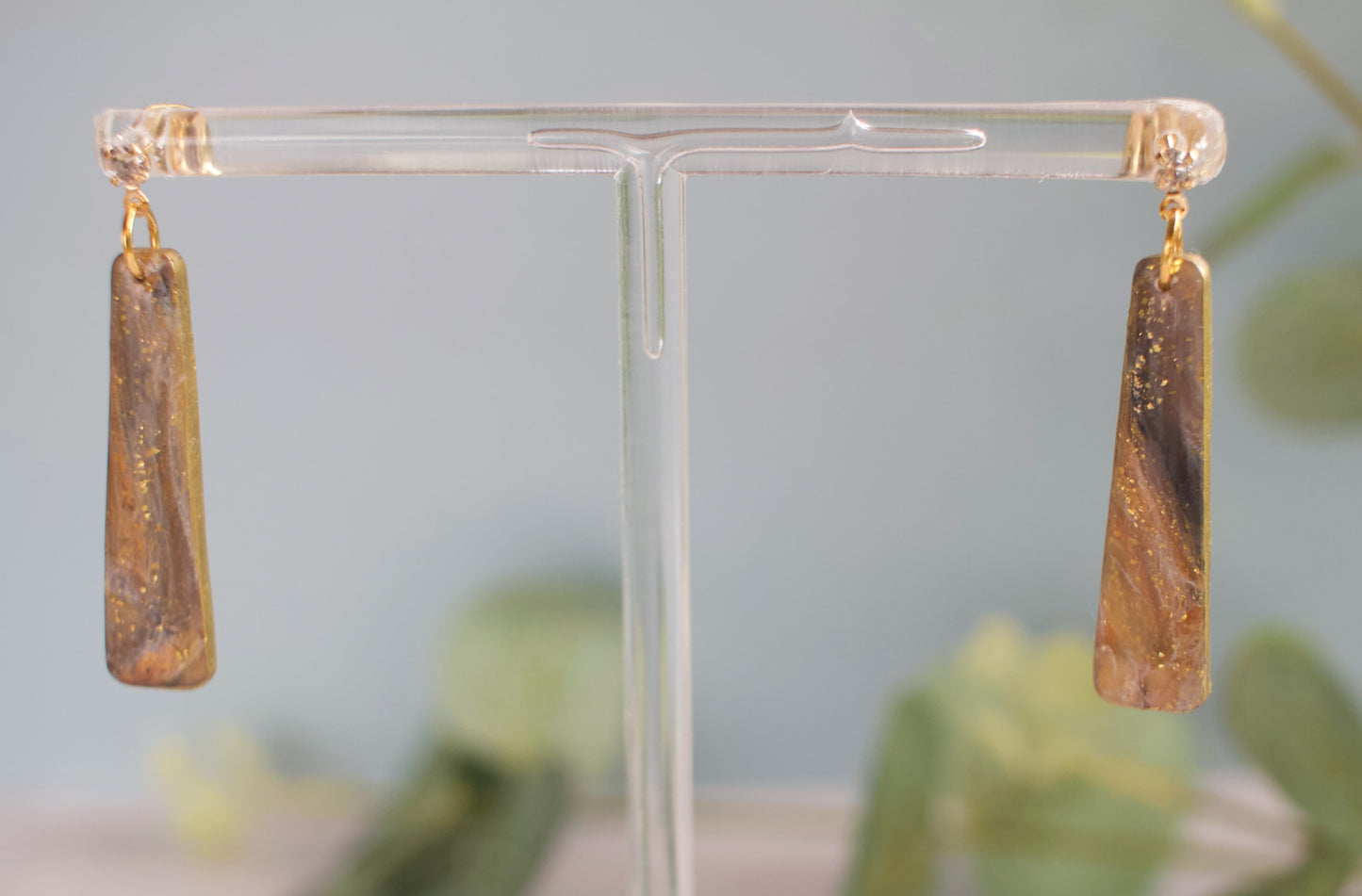 Gold and black marbled long earrings on a clear stand with a blurred green background