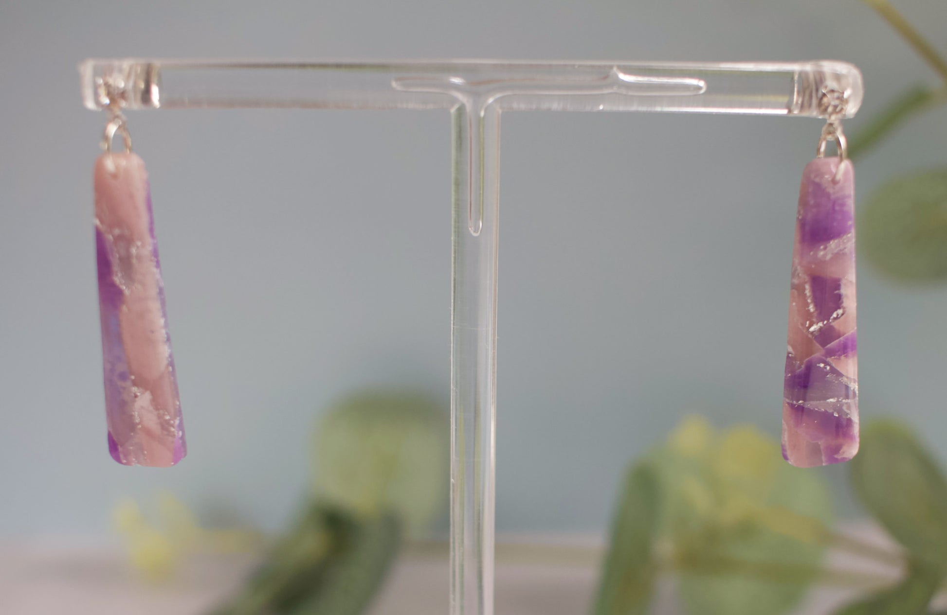Pink and purple marbled earrings on a clear stand with a blurred green plant background