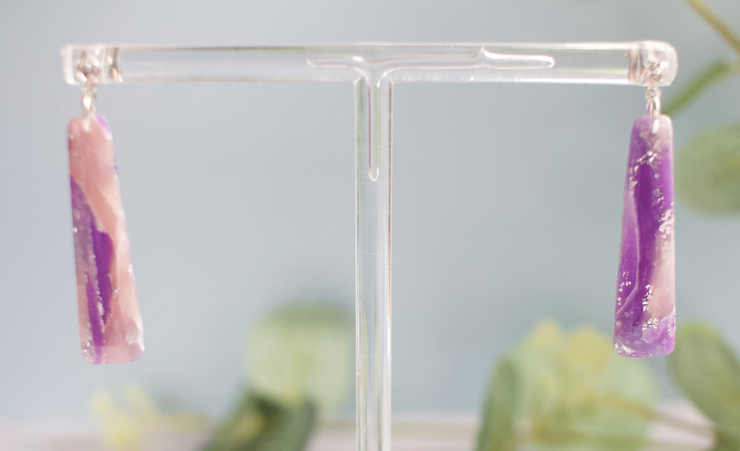 Pair of pink and purple bar shaped earrings on a clear stand with a blurred green plant background.