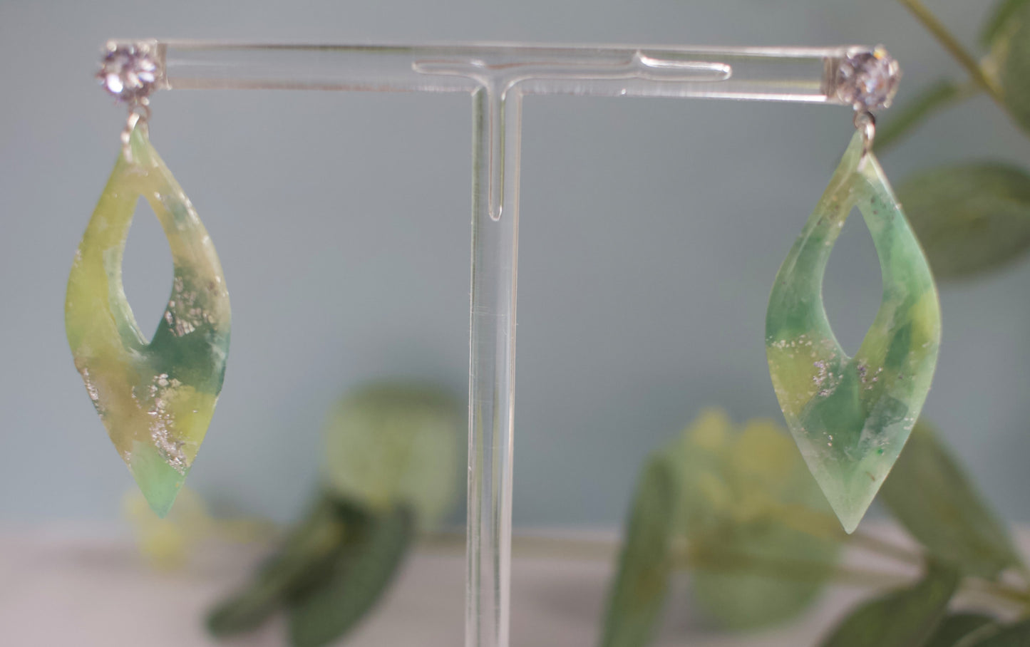 Green leaf-shaped earrings on a clear stand with a blurred green background
