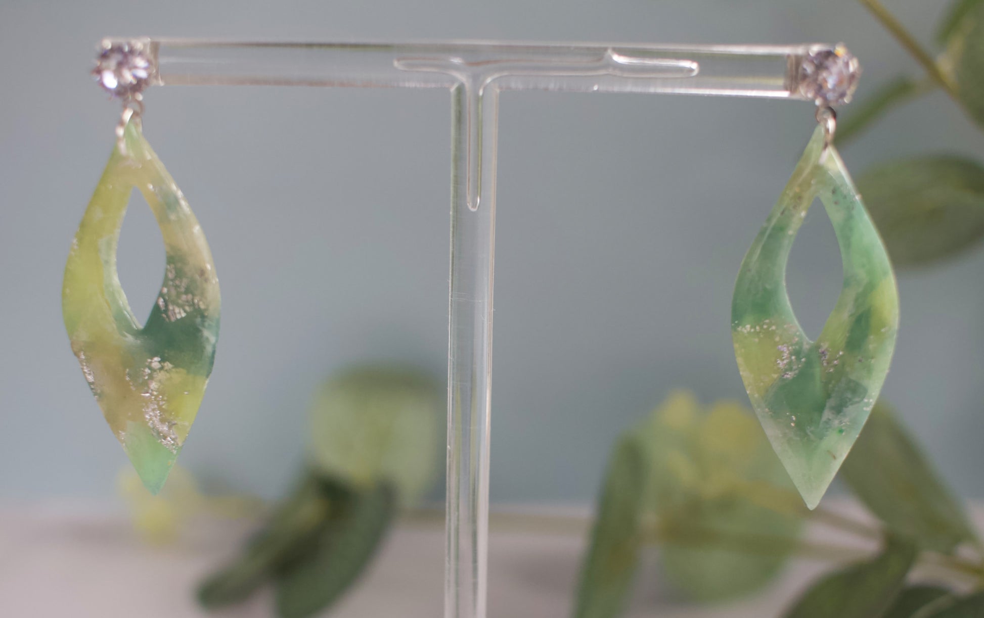 Green leaf-shaped earrings on a clear stand with a blurred green background