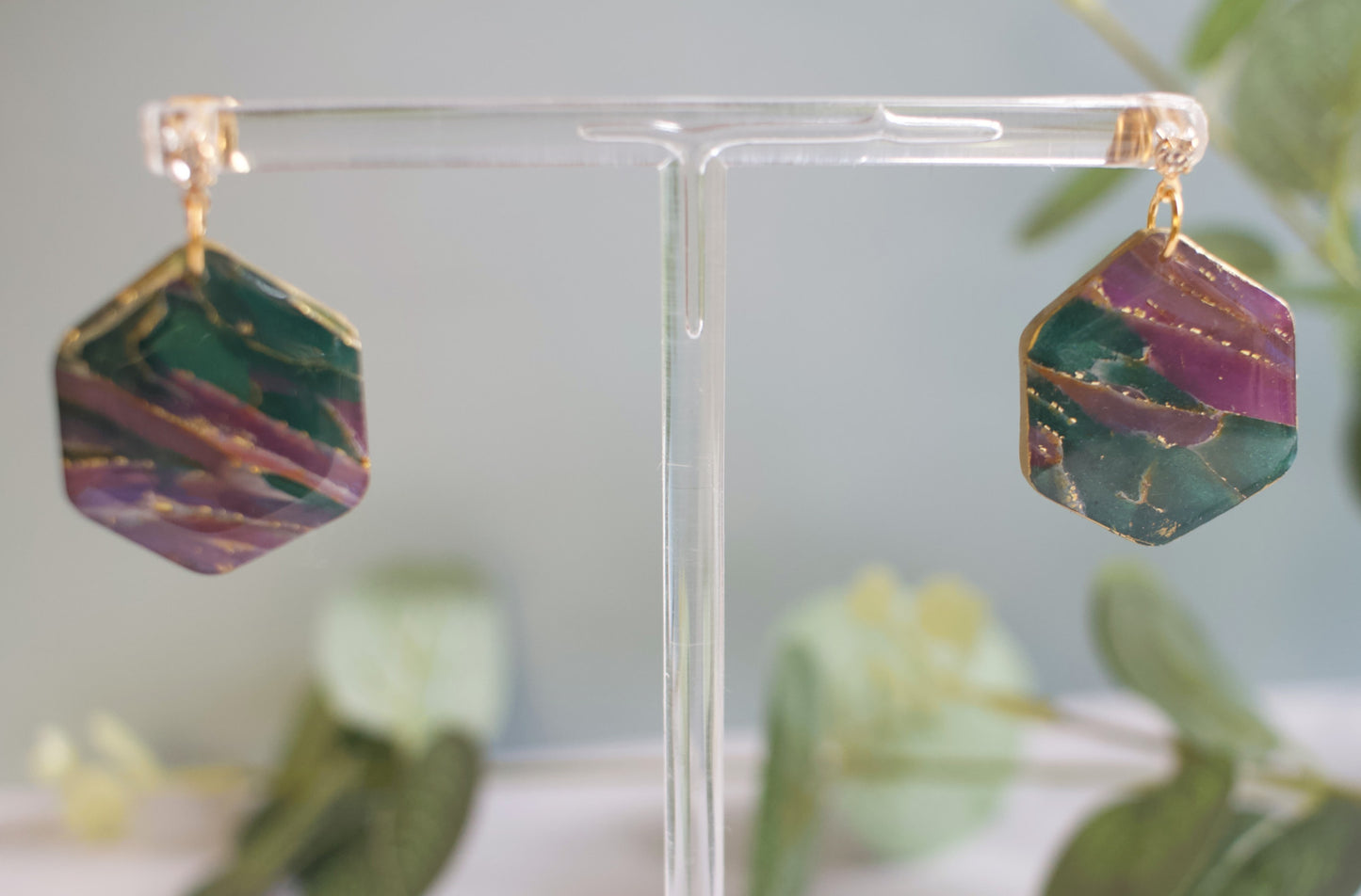 Hexagonal earrings with green and purple marbled design on a clear stand with a blurred plant background.