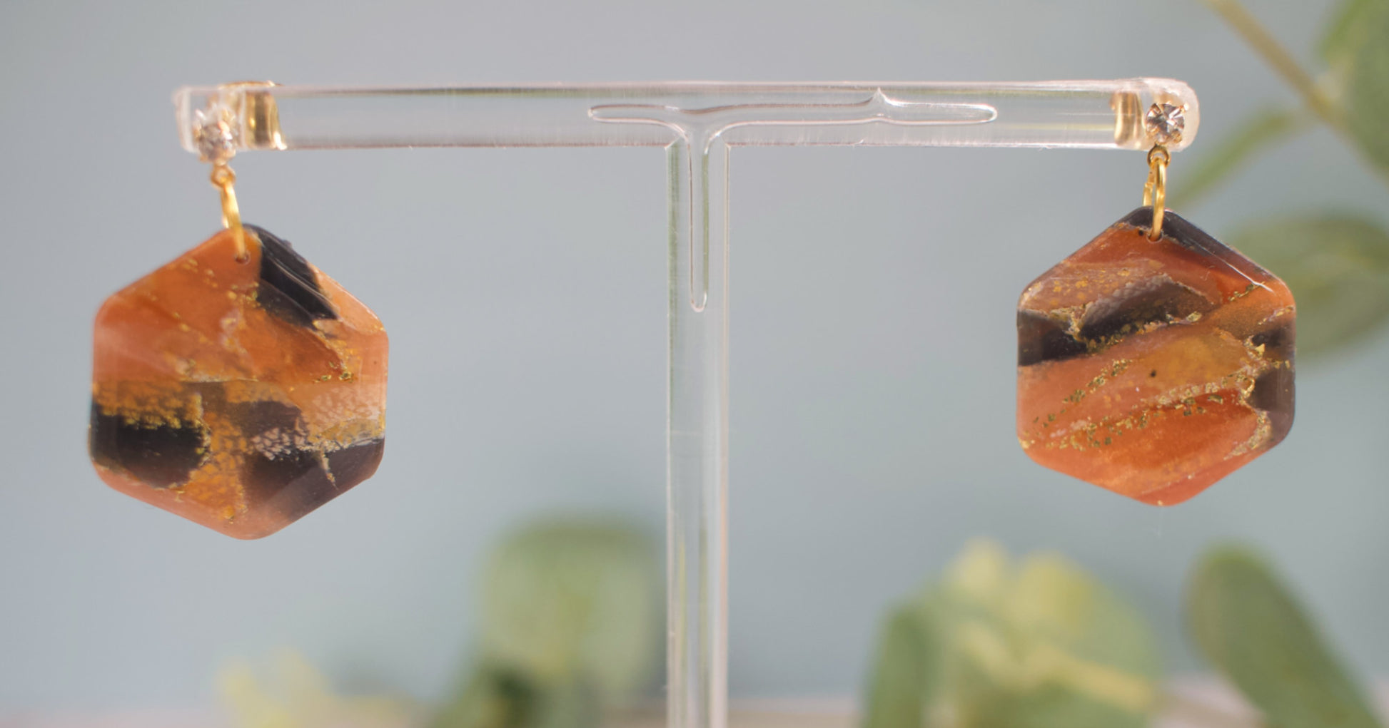 Hexagonal orange and brown marbled earrings on a clear stand with a blurred green plant background