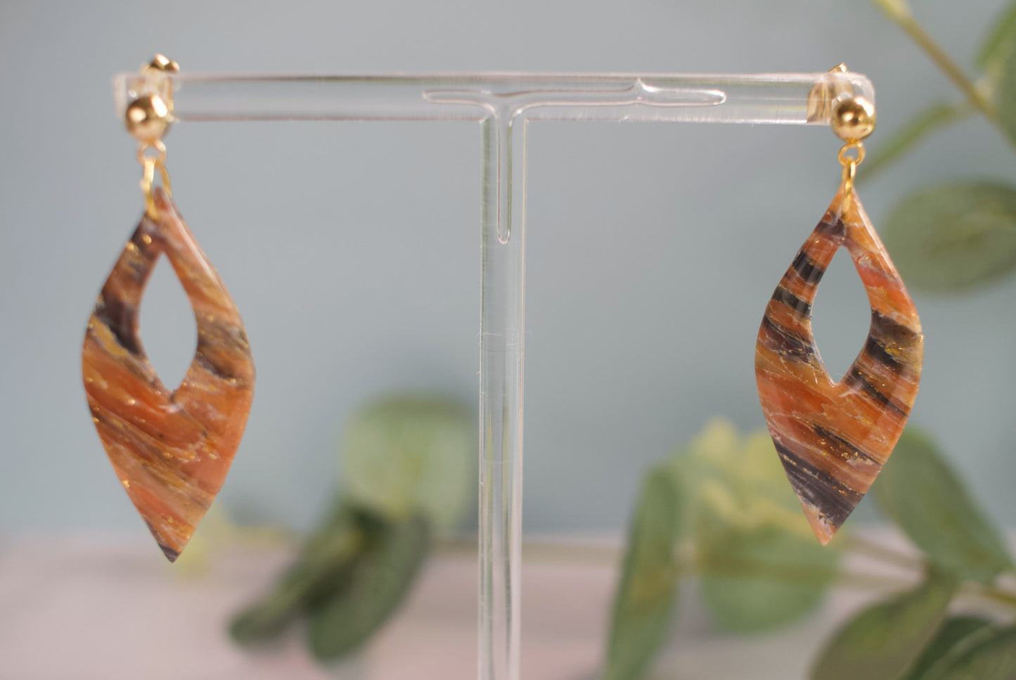 Pair of orange and gold earrings on a clear stand with a blurred green plant background