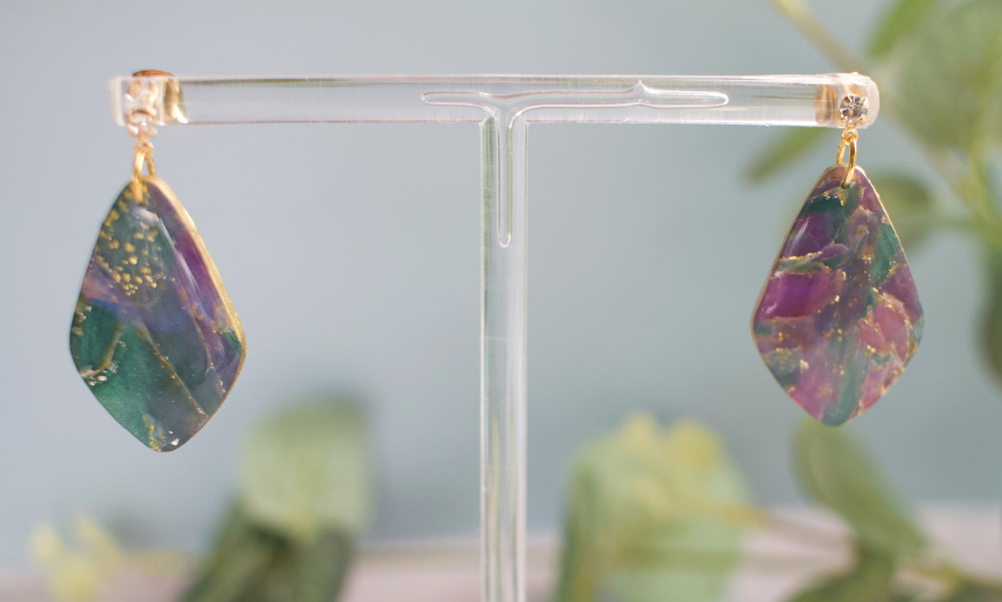 Teardrop-shaped purple and teal marbled earrings on a clear stand with a blurred green plant background