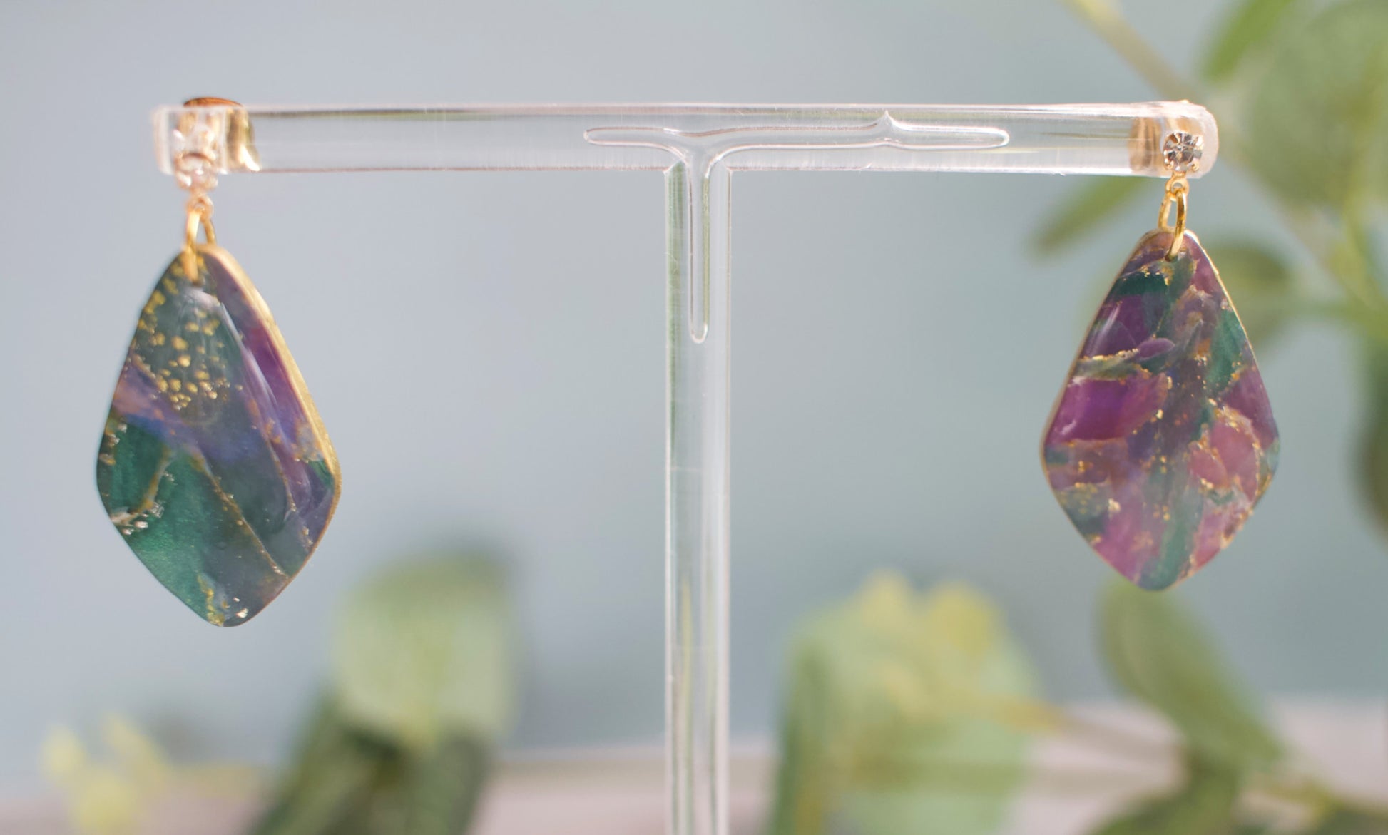 Teardrop-shaped purple and teal marbled earrings on a clear stand with a blurred green plant background