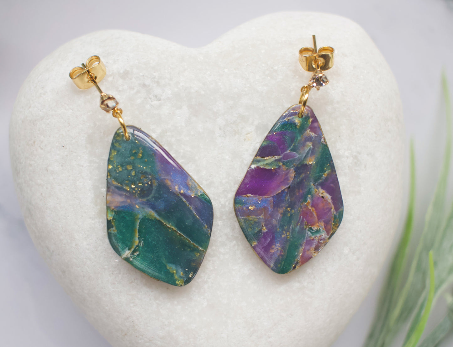 Purple and teal marbled earrings on a heart-shaped white stone with a blurred green plant in the background