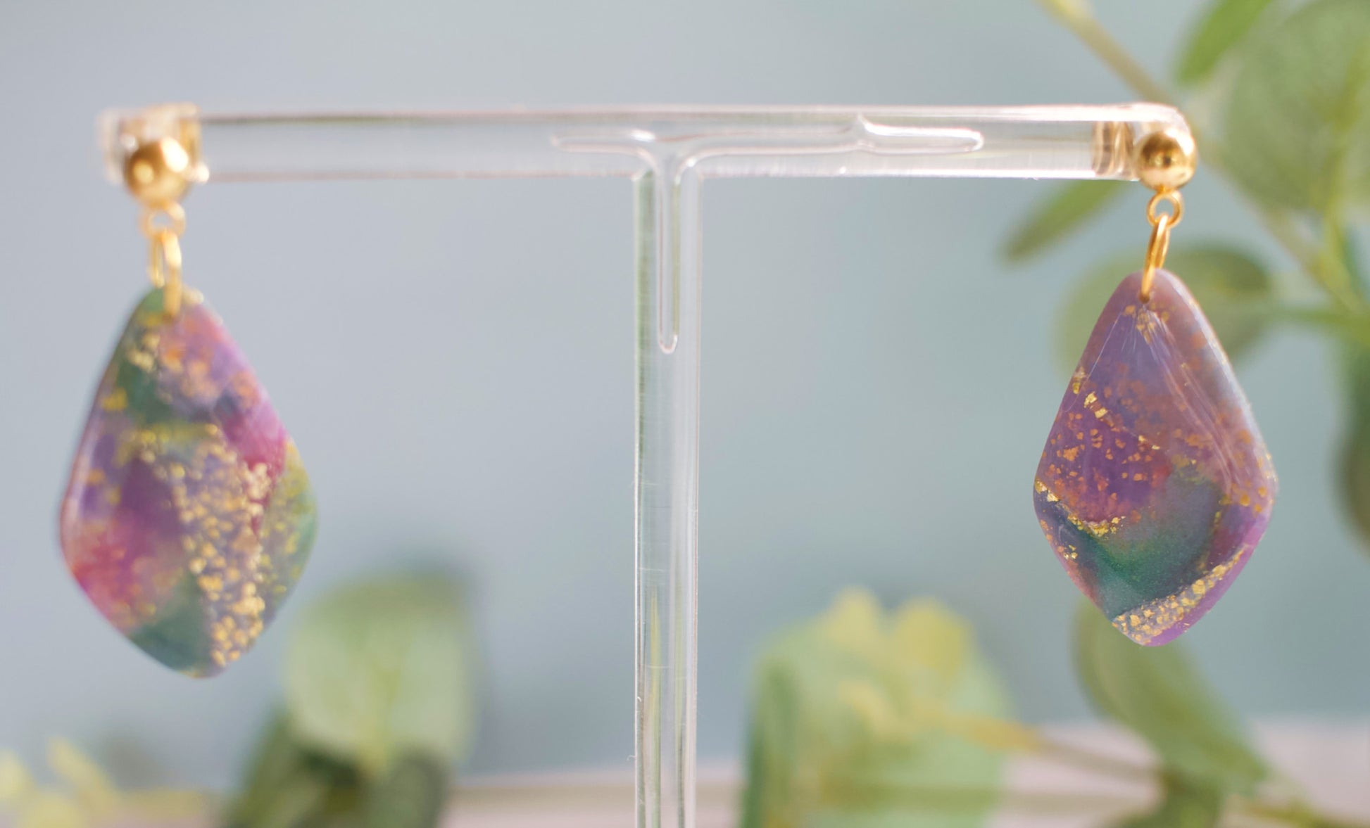Teardrop-shaped earrings with purple and teal marbled design on a clear stand against a light blue background.