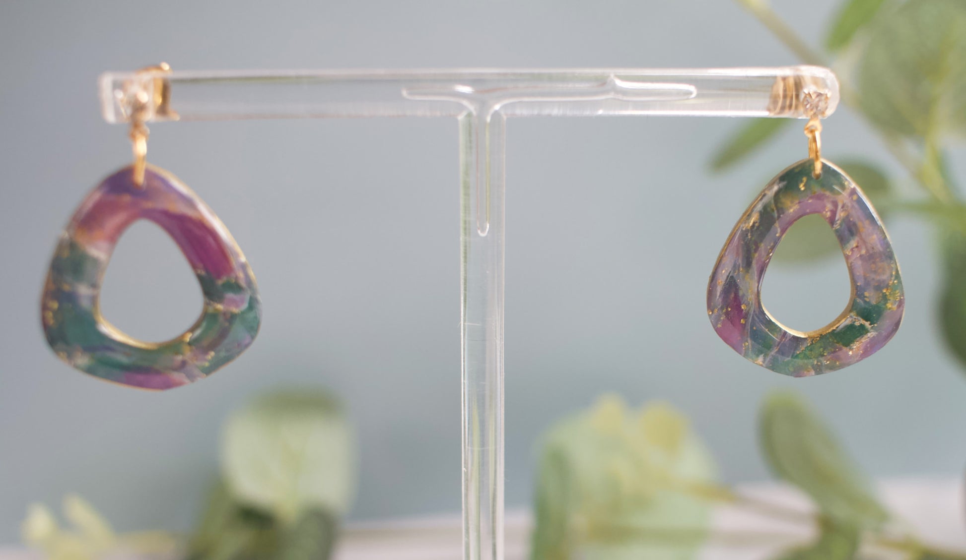 Purple, teal and gold teardrop-shaped earrings on a clear stand with a blurred green background