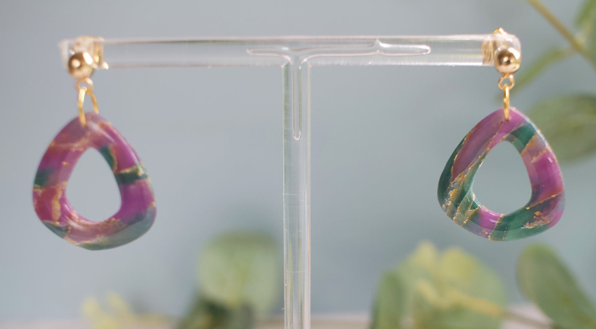 Purple and teal marbled teardrop-shaped earrings on a clear stand with a blurred green background