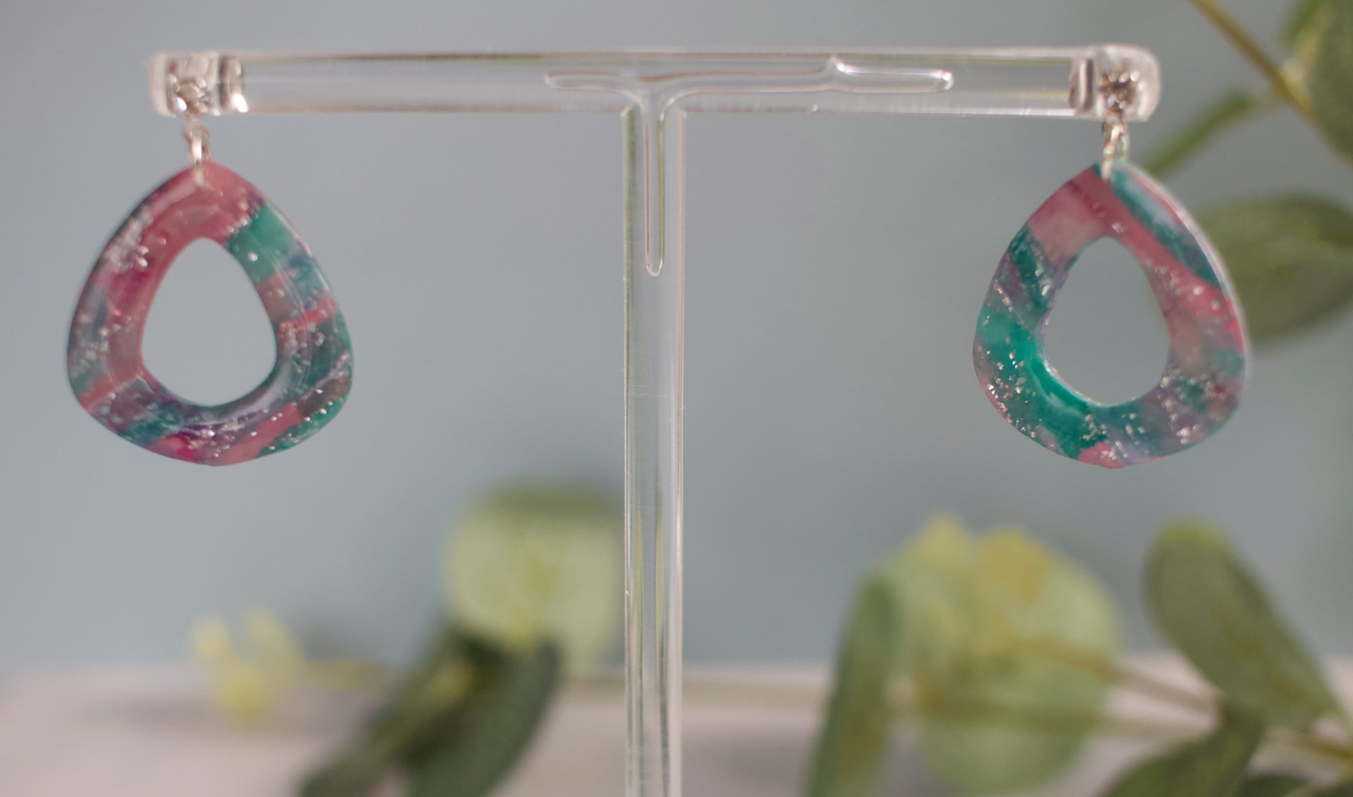 Colorful teardrop-shaped earrings on a clear stand with a blurred green plant background