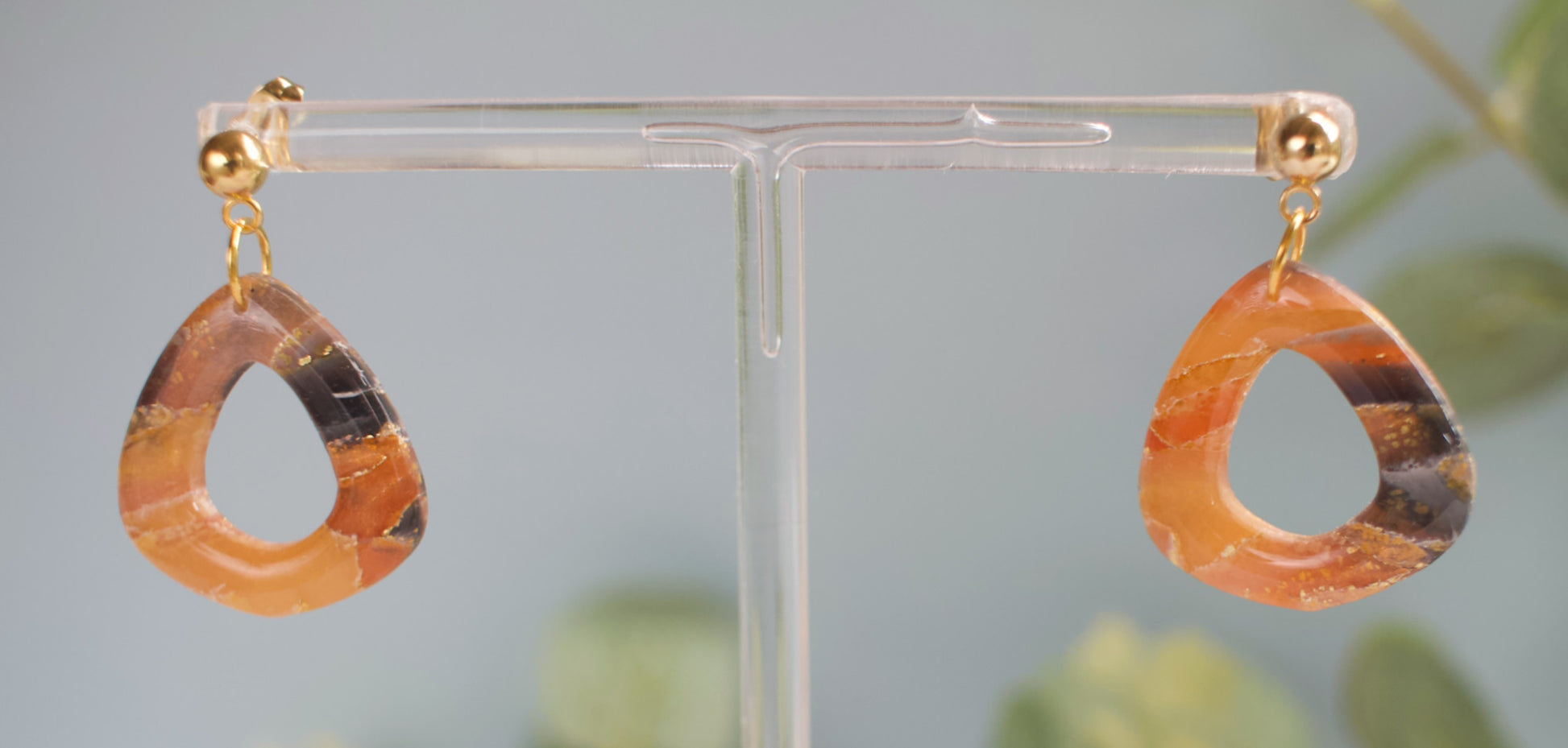 Teardrop-shaped earrings with gold hooks on a clear stand against a blurred natural background.