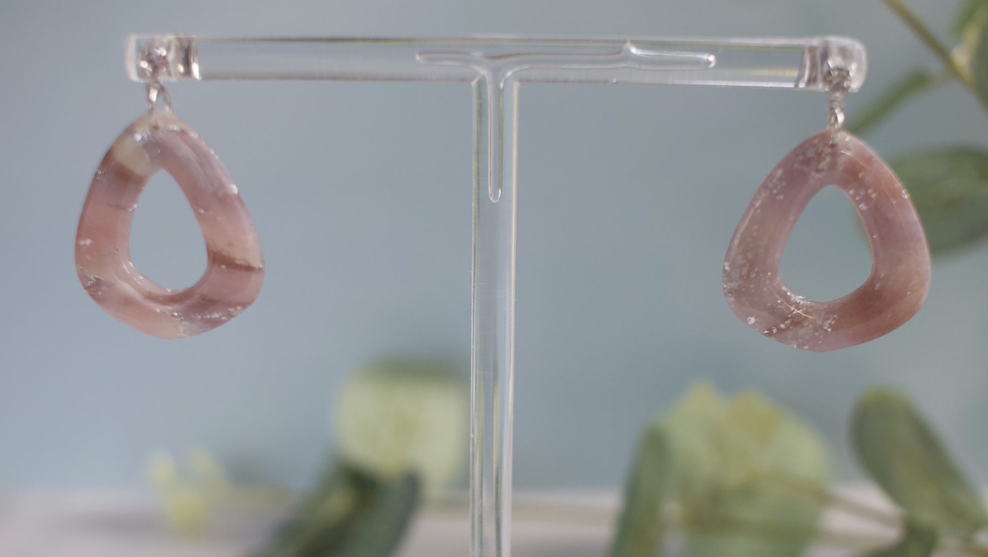 Pink teardrop earrings on a clear stand with a blurred green plant background