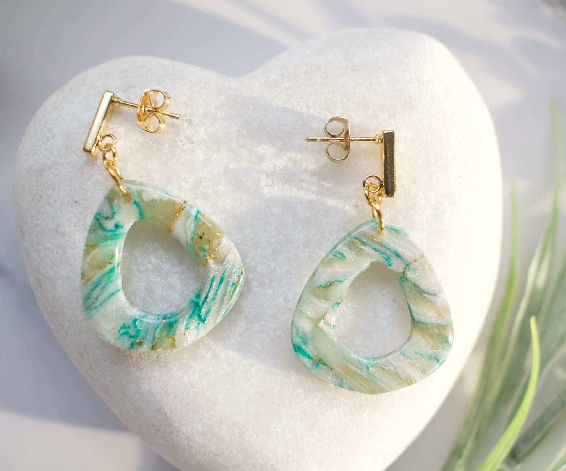 Marble-patterned earrings on a white heart-shaped stone with a blurred green plant in the background.