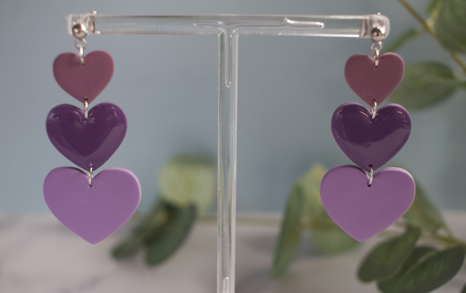 Purple heart-shaped earrings on a clear stand with a blurred green plant background